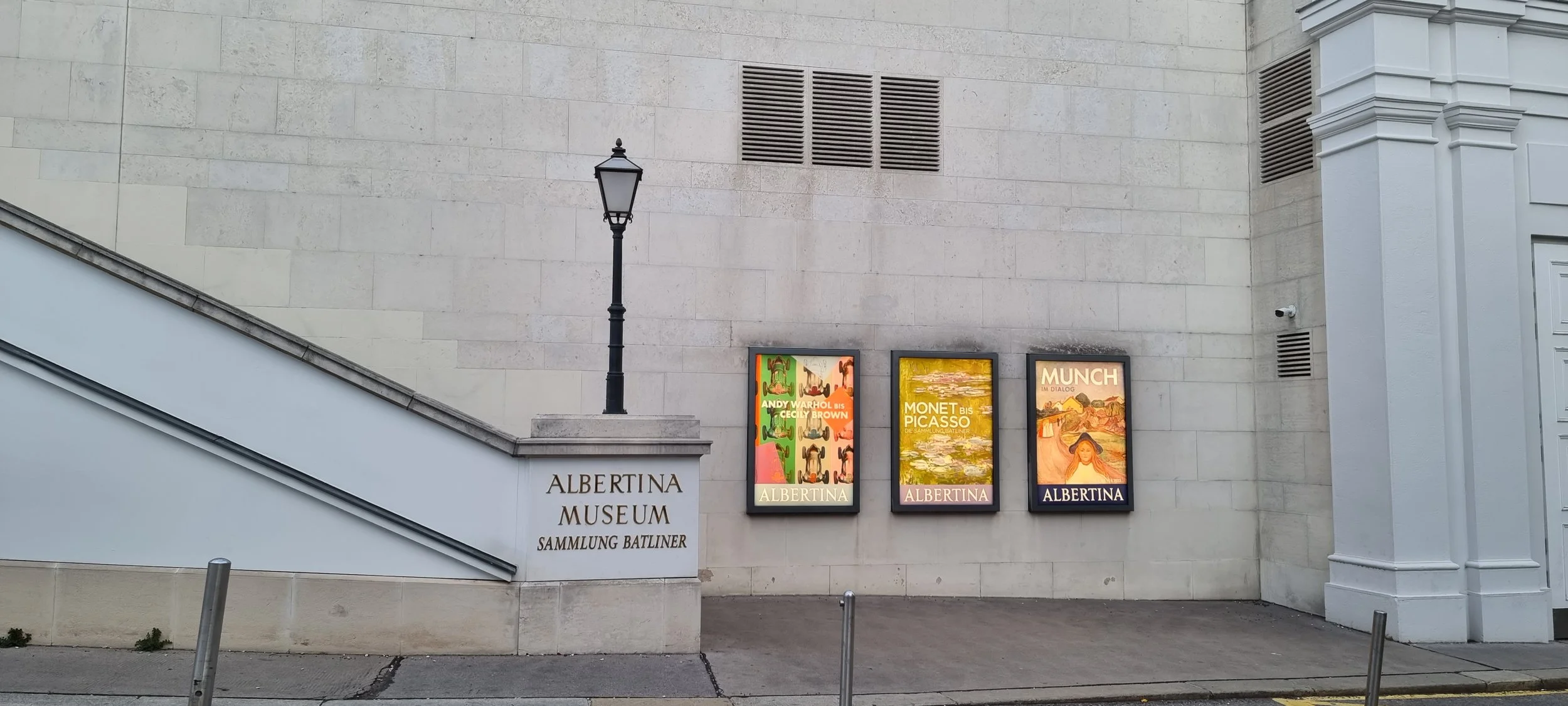 Exterior view of the Albertina Museum in Vienna, Austria, displaying illuminated posters of art exhibitions, including works by Andy Warhol, Monet, Picasso, and Munch, on a large beige stone wall with a streetlamp nearby.