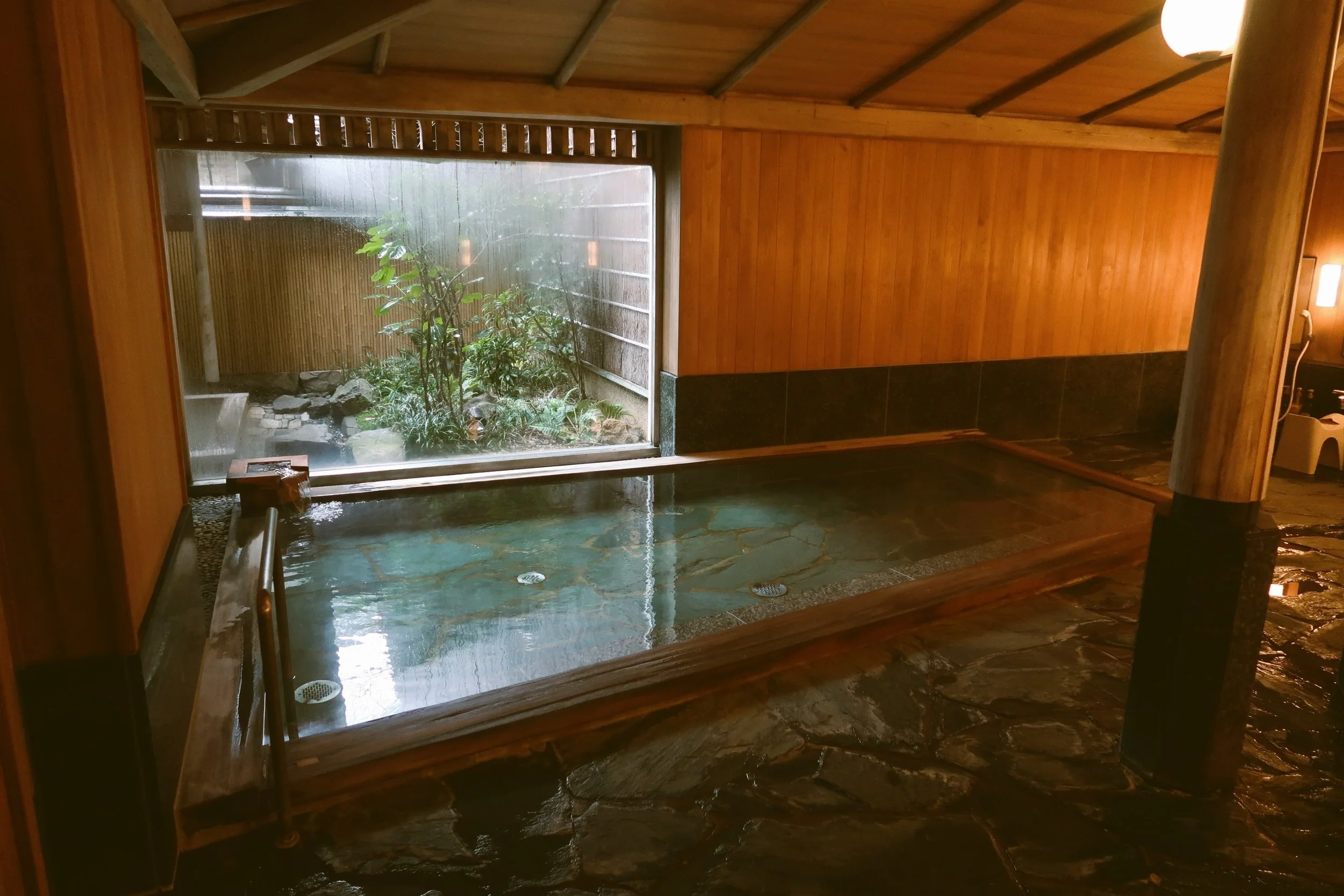 Indoor onsen with a wooden-framed bath and a large window looking onto a garden, Japan.