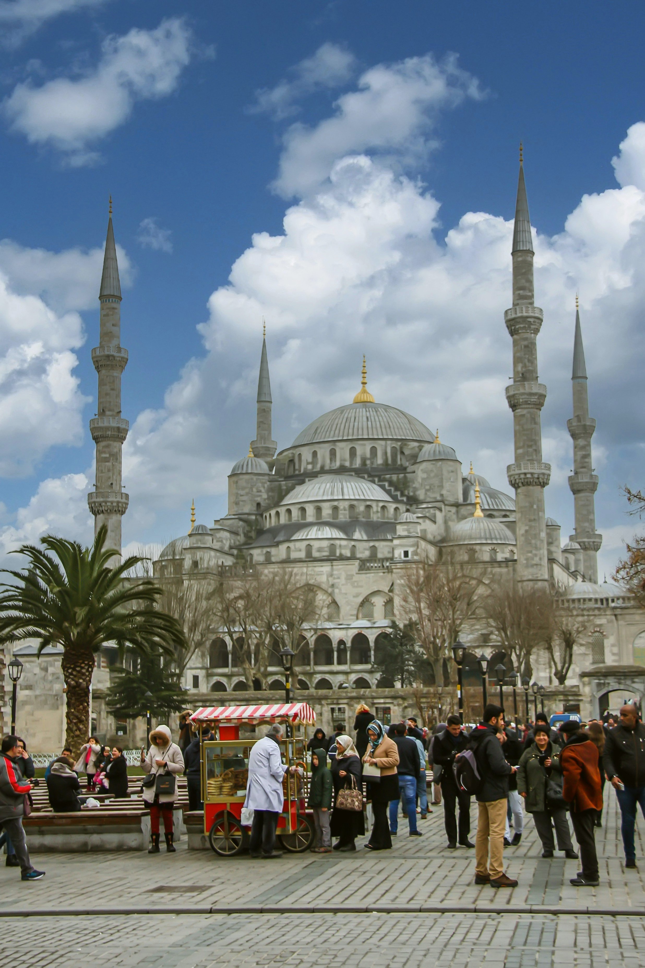 Blue Mosque with its domes and six minarets under a blue sky with clouds, and visitors in the square in front, Istanbul.