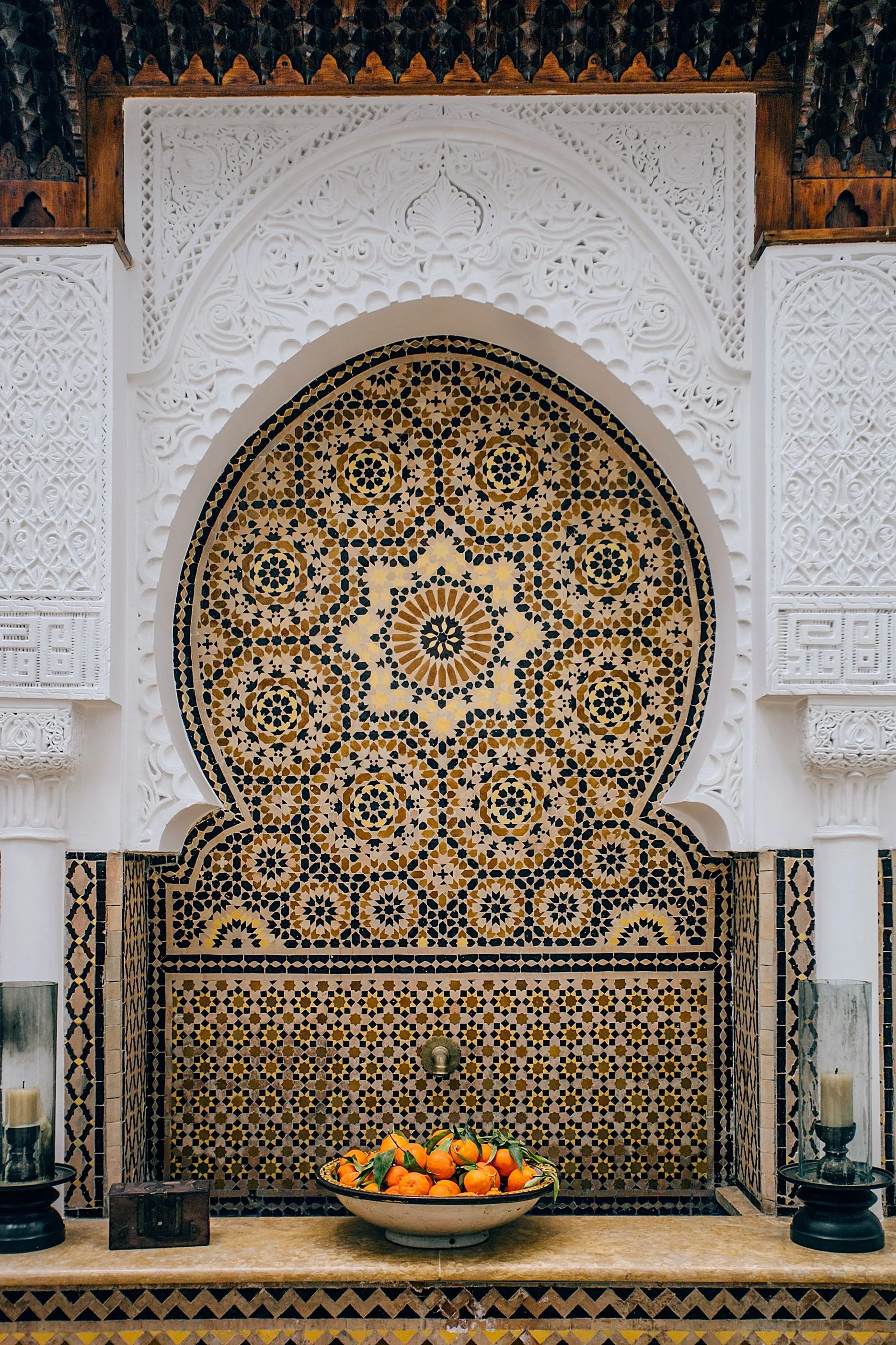An ornate altar area featuring intricate white carved designs and mosaic tiles with a geometric pattern in warm earth tones. A bowl of oranges is placed on the altar, flanked by candles and decorative items.