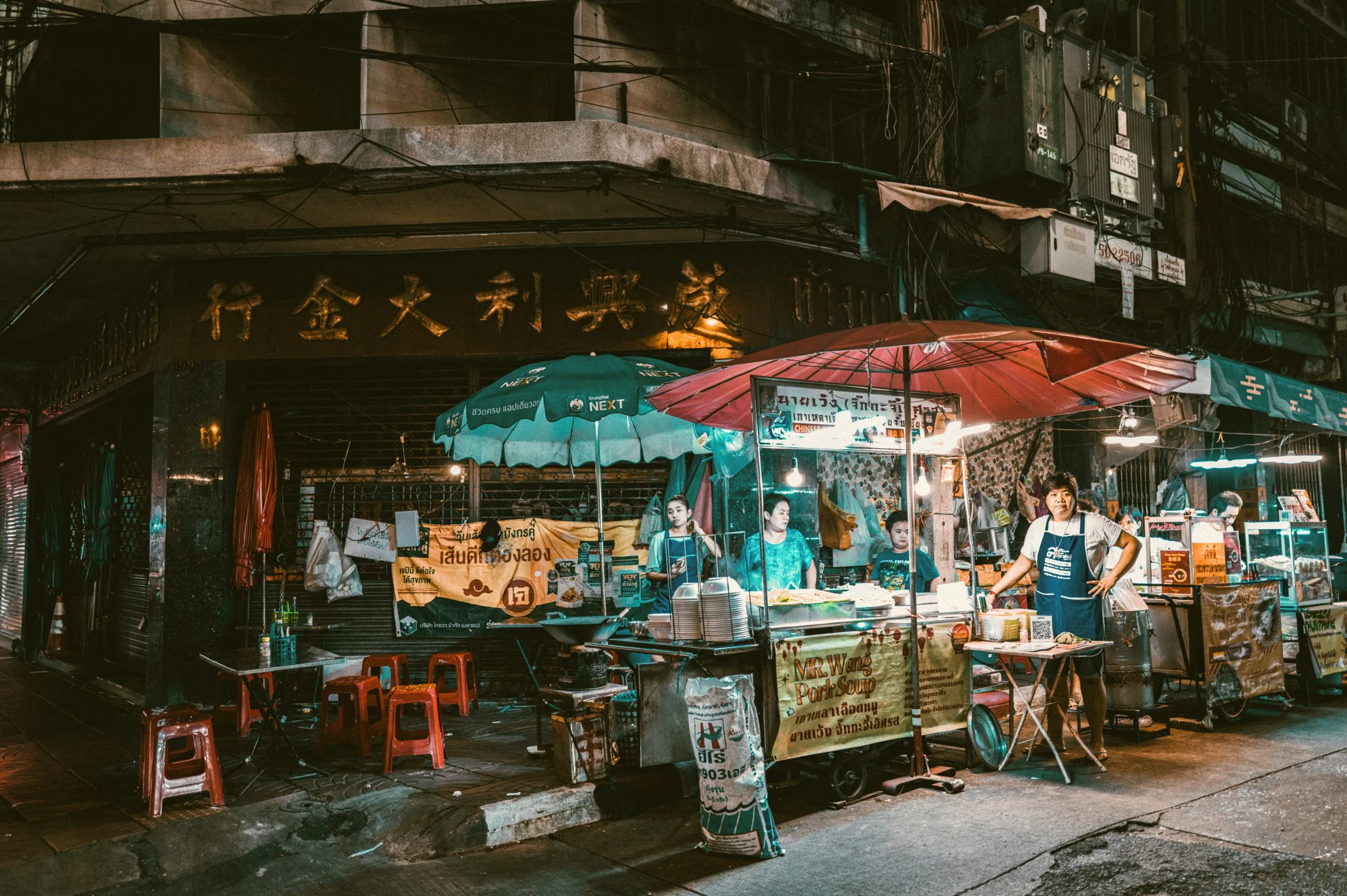 Street food stalls with colorful umbrellas lit up at night on a dark street, Bangkok