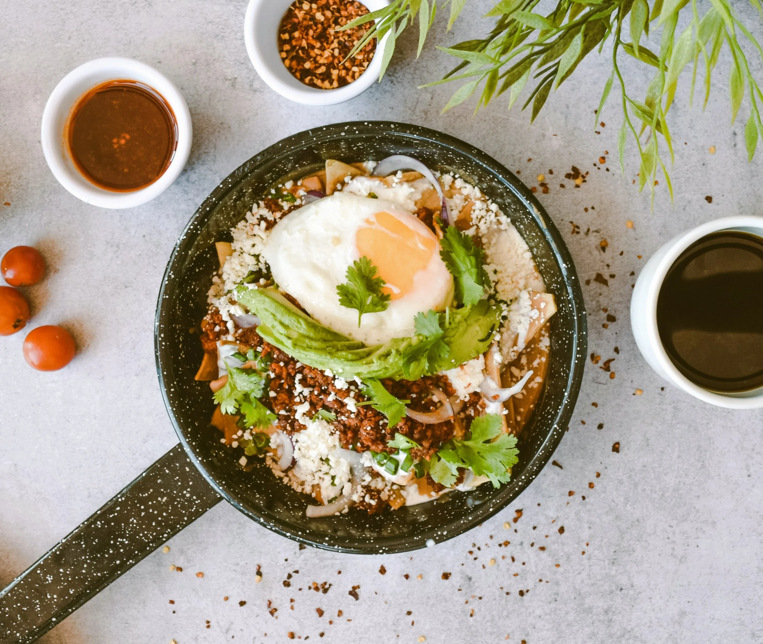 Skillet of chilaquiles topped with a fried egg and avocado, served with small bowls of salsa and a cup of coffee, Mexico City.