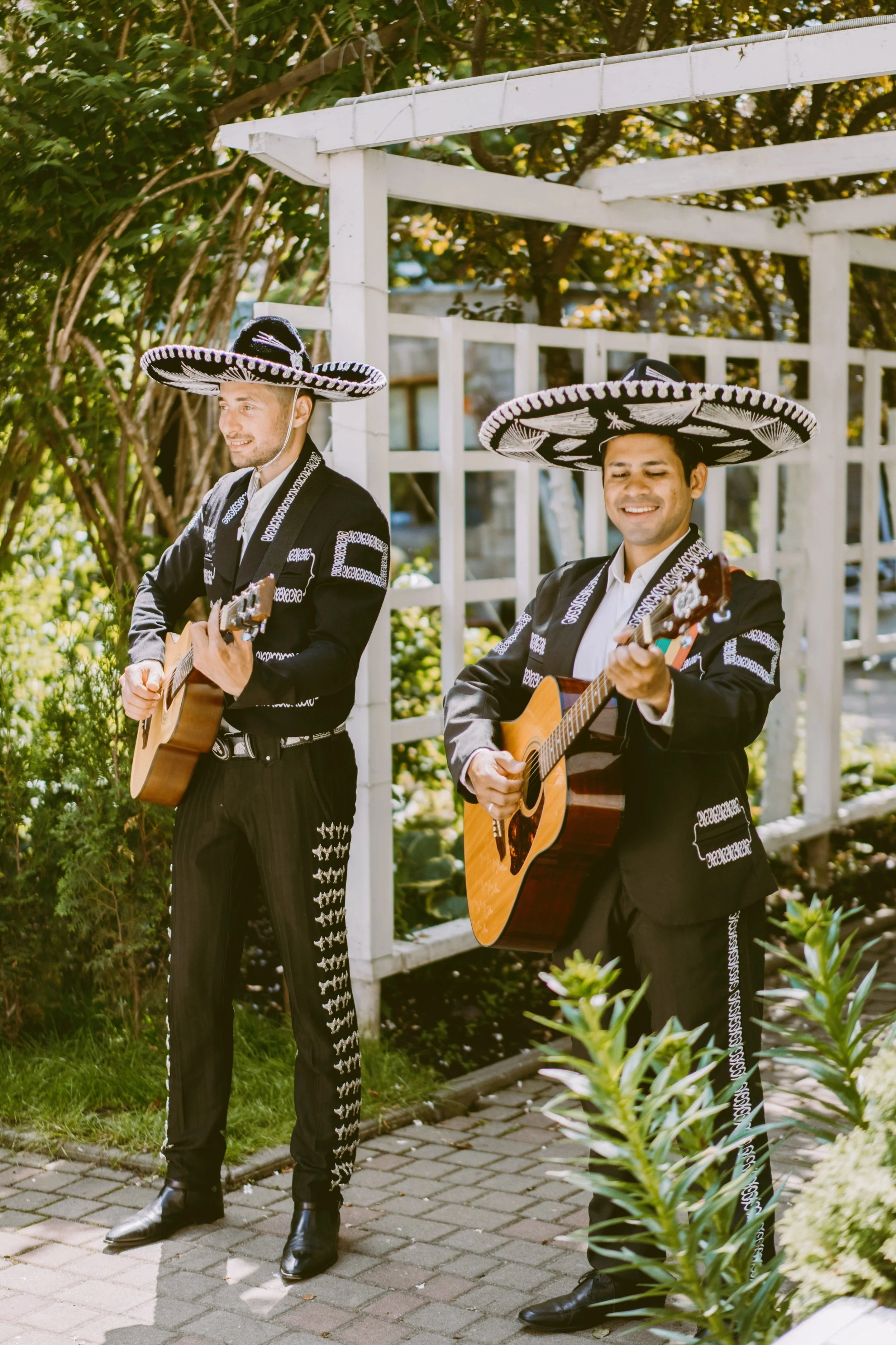Two mariachi musicians in charro outfits playing guitars in front of a white pergola surrounded by greenery, Mexico City.