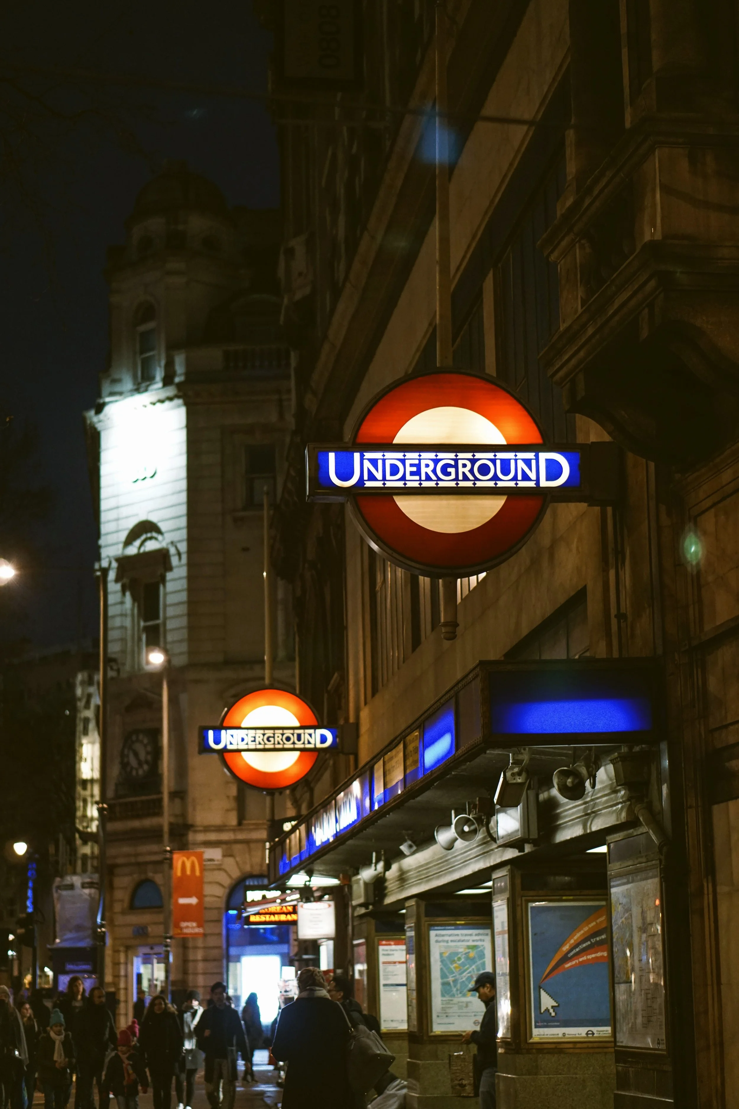 London Underground sign at night with street scene filled with people and illuminated buildings.