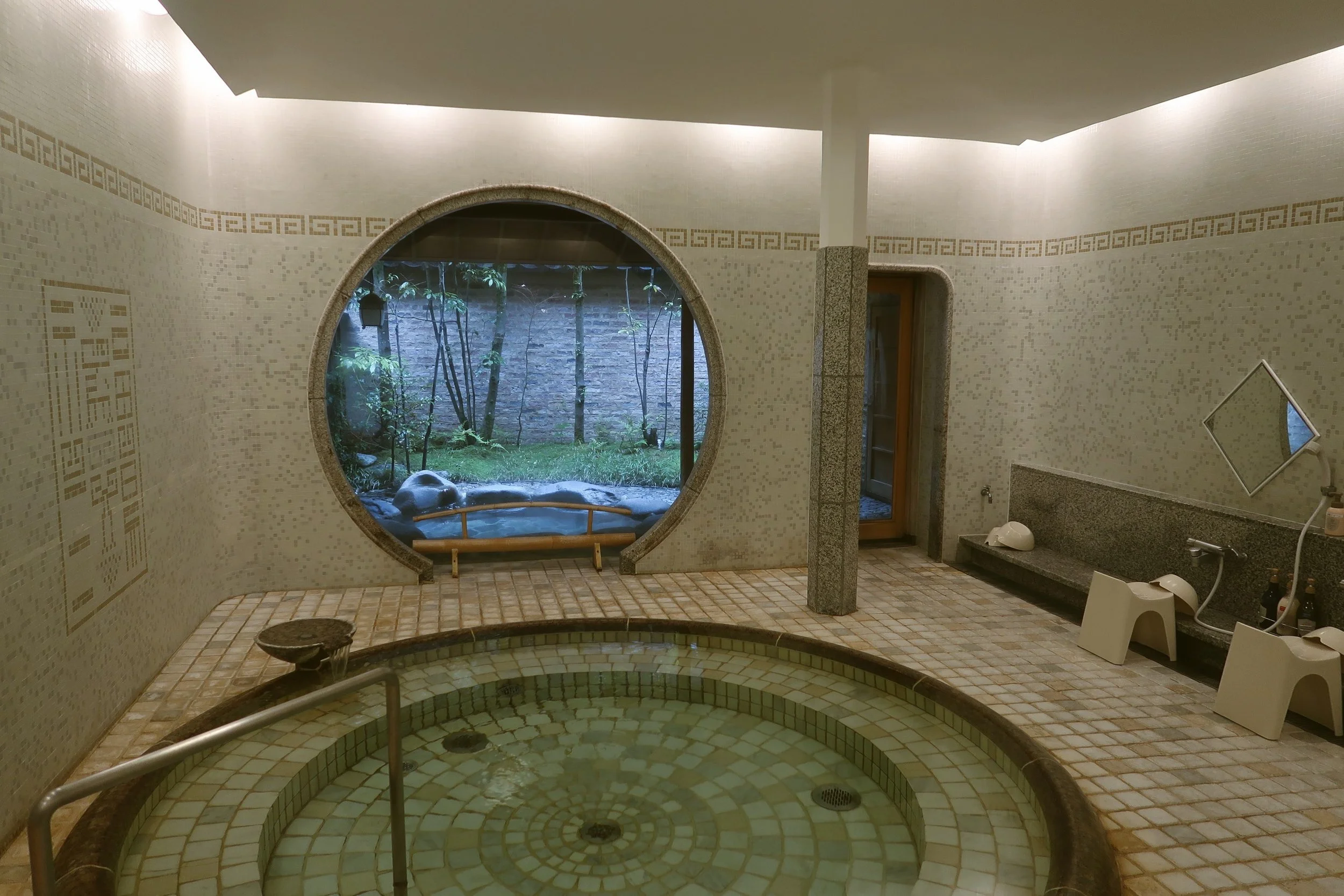 Circular onsen pool beneath a round window overlooking greenery, in a bright stone-walled room, Japan.