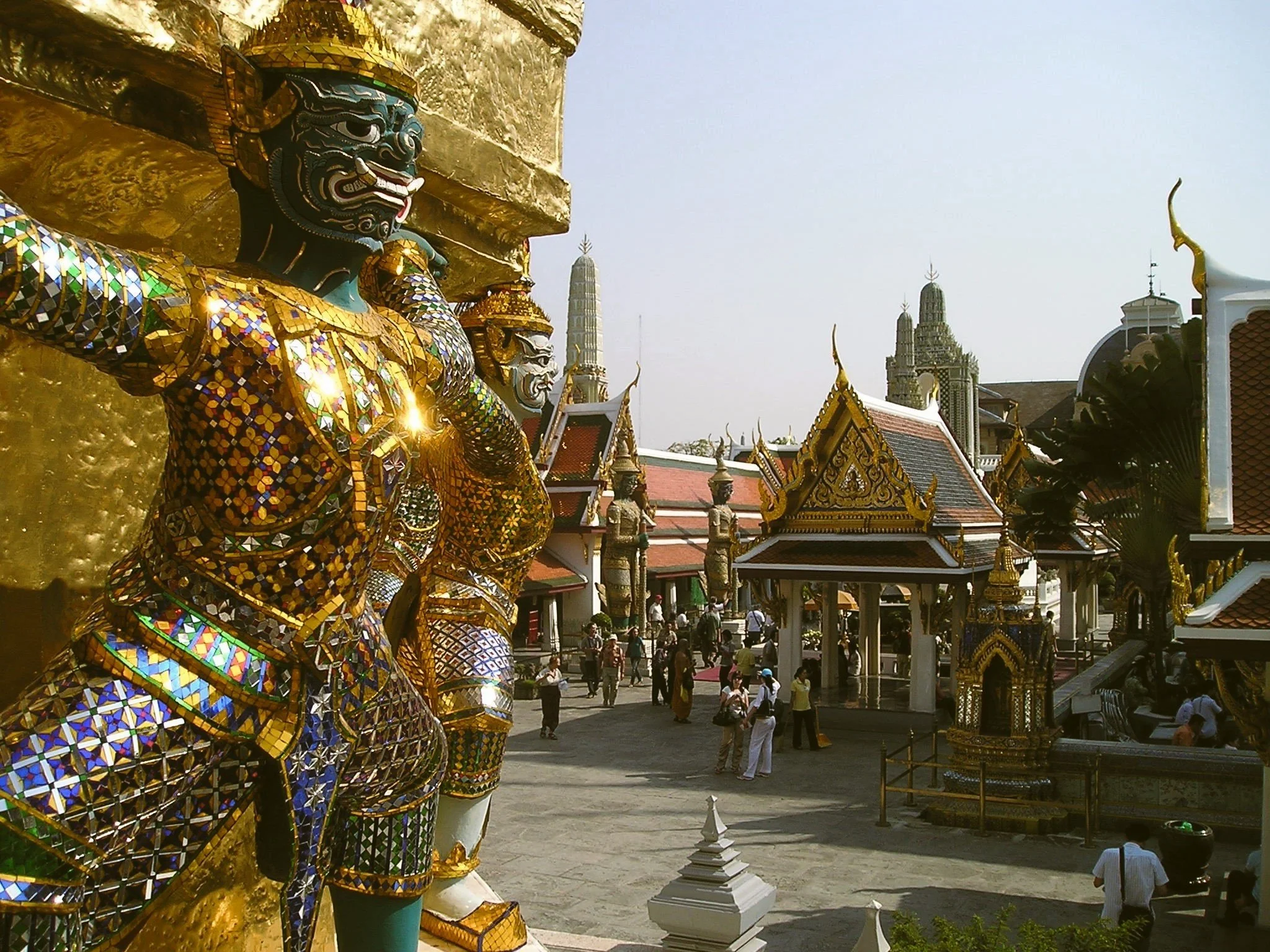 Golden mosaic-covered guardian statue in the foreground with temple roofs behind it, Grand Palace, Bangkok.