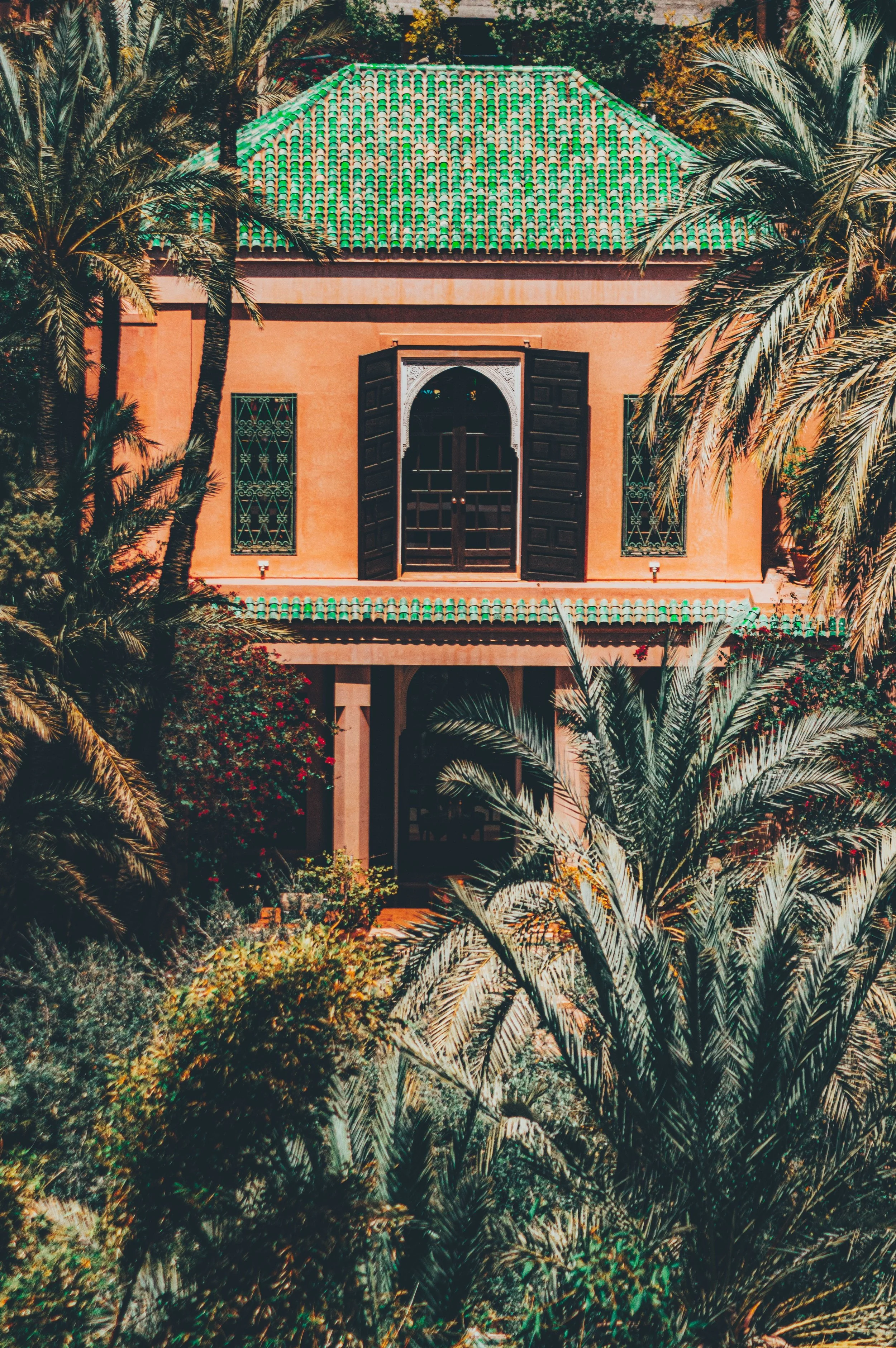 Coral-colored building with a green tiled roof among dense palm fronds, Jardin Majorelle, Marrakech.