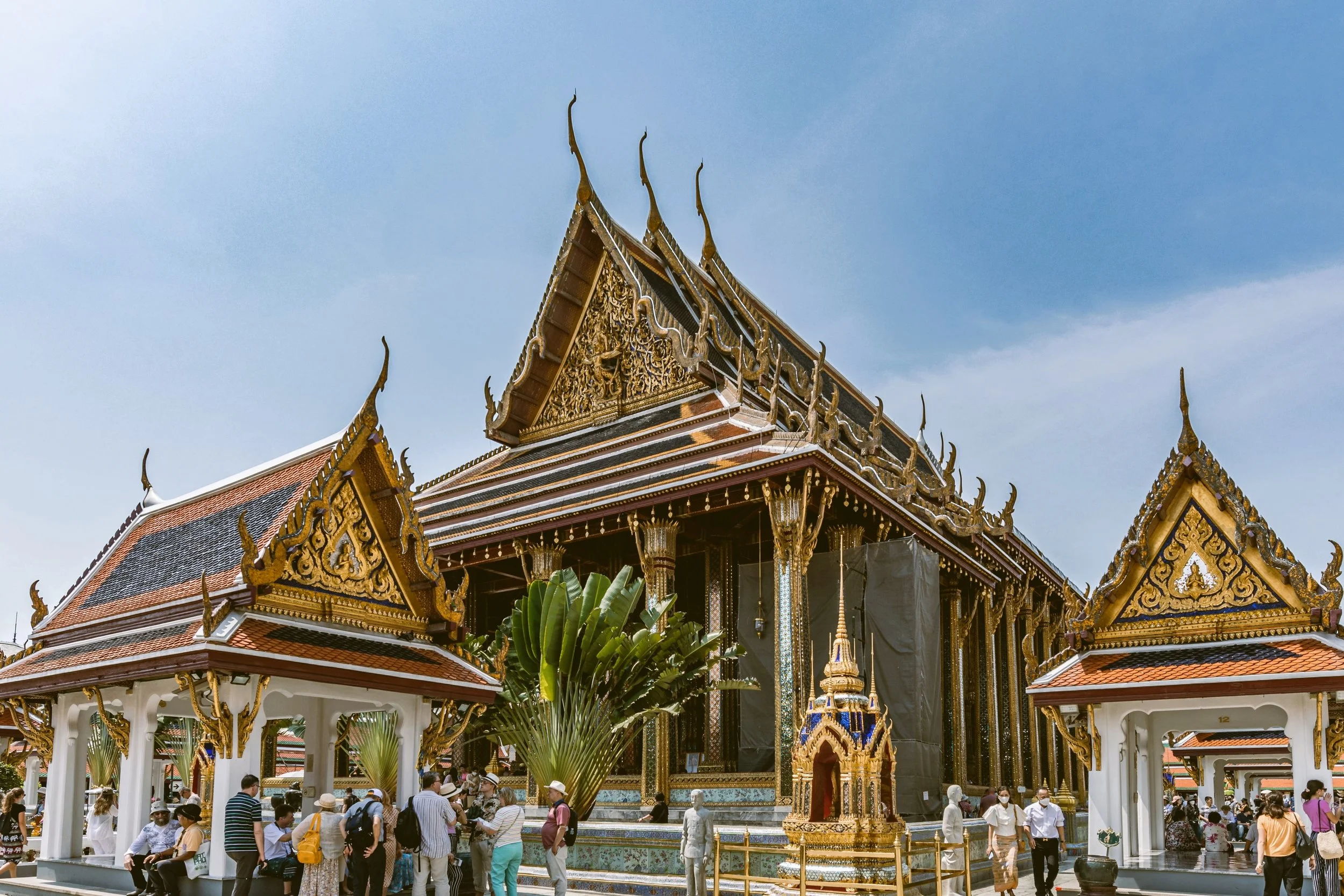 Temple with a tiered roof and golden decorations, with visitors gathered in front under a blue sky, Grand Palace, Bangkok.
