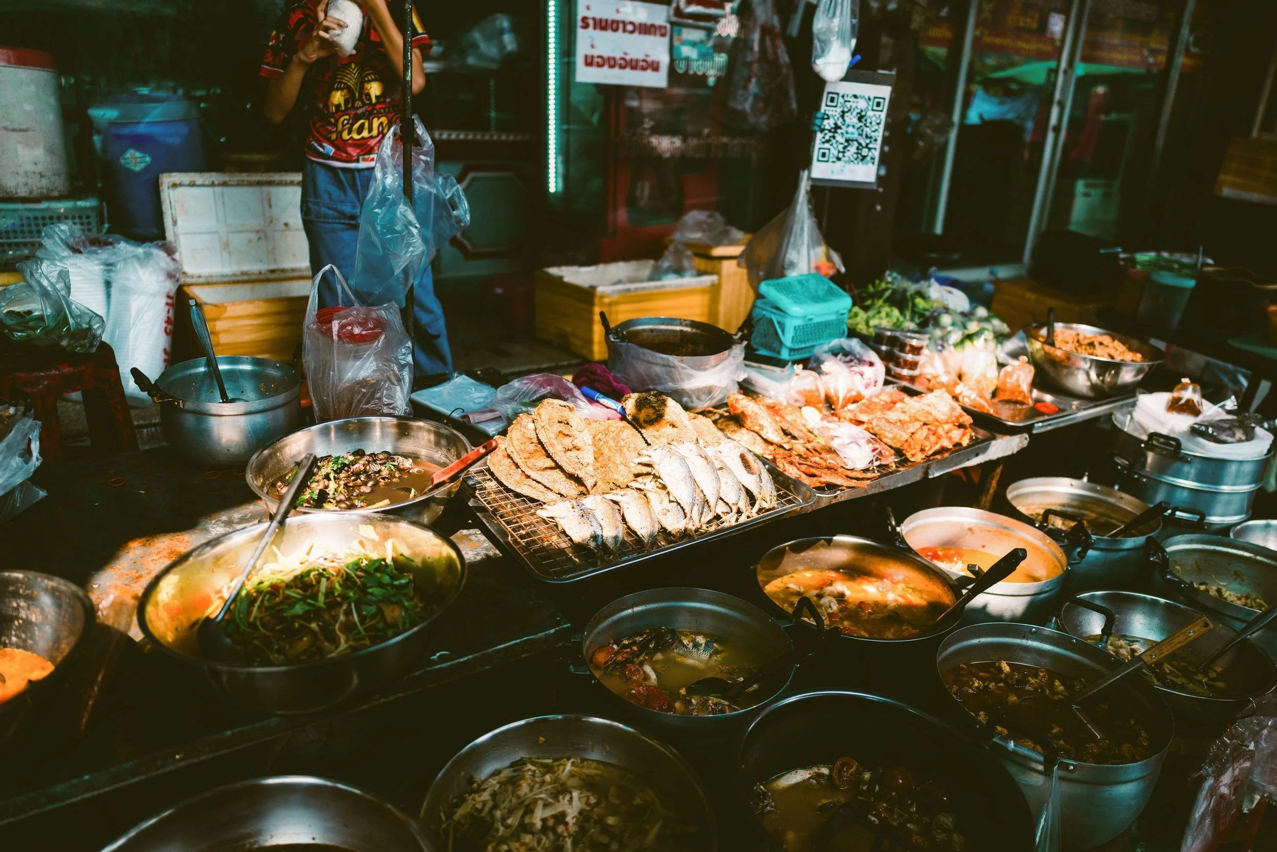 Street food stall at night with trays of cooked dishes and a vendor behind the counter, Bangkok.