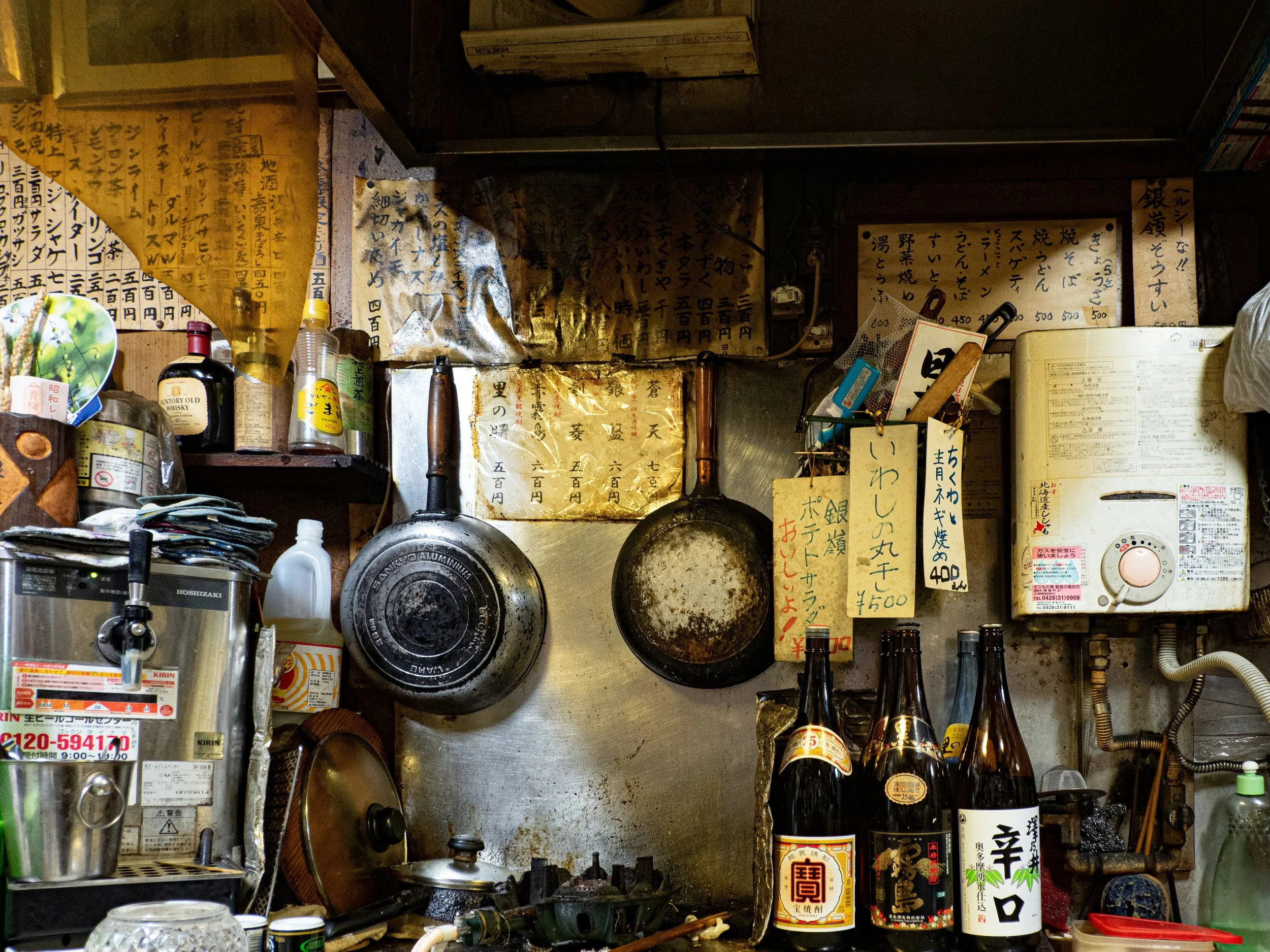 Cluttered izakaya kitchen with hanging pans, utensils and sake bottles on a shelf, Tokyo.