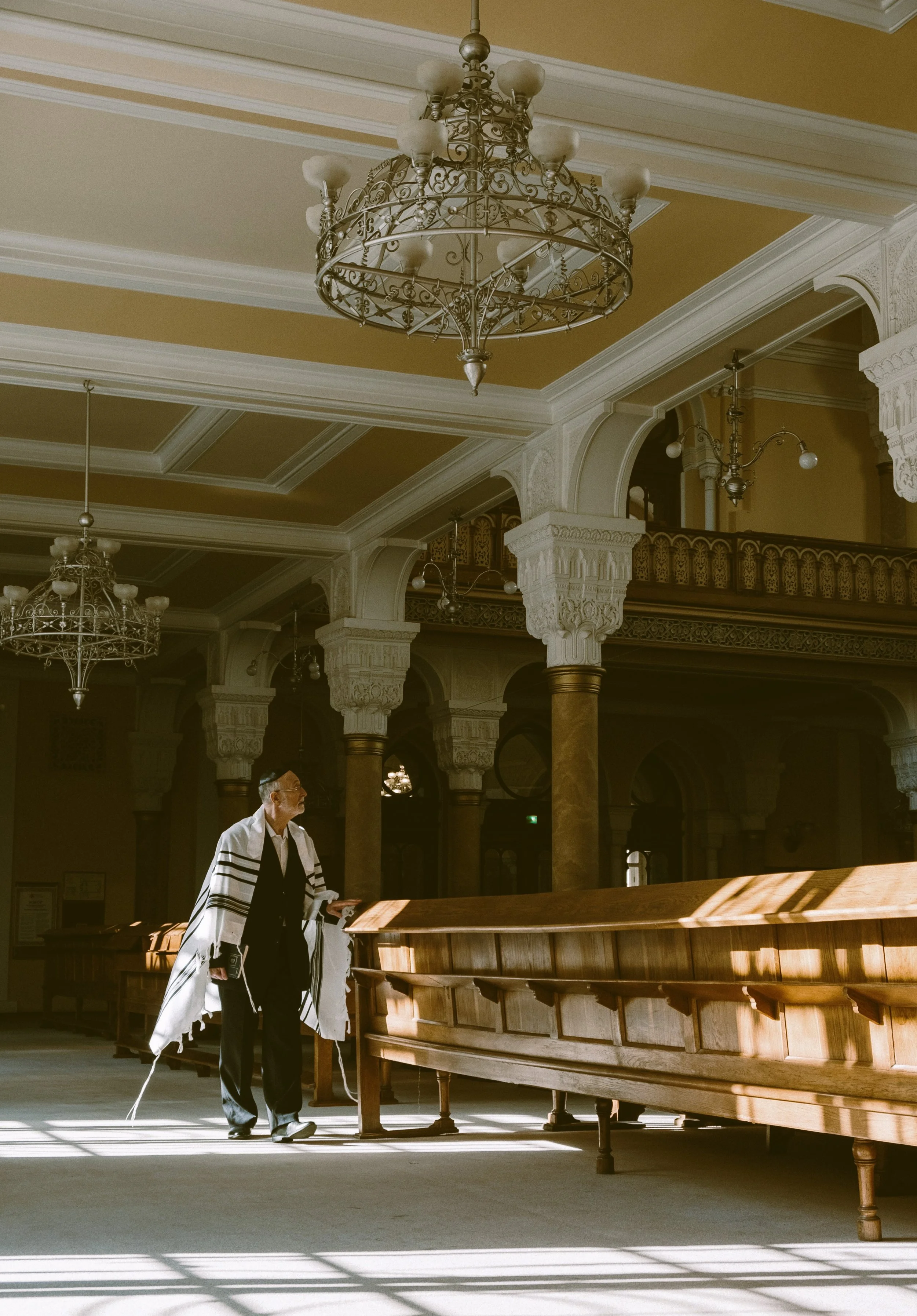 Man wearing a tallit praying at a wooden ark in a synagogue with ornate chandeliers and decorative columns, Budapest