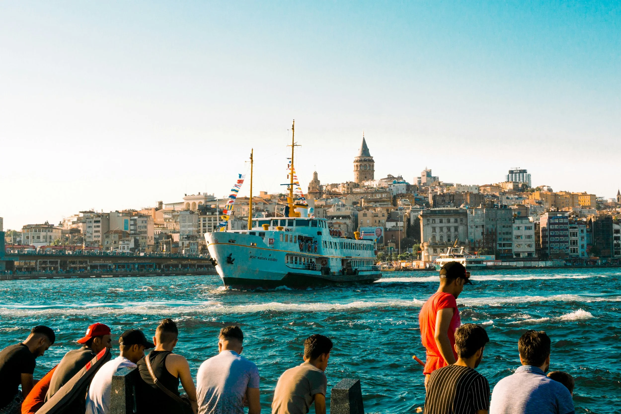 People sitting along the shore watching a ferry cross the Bosphorus, with the Galata skyline behind, Istanbul.