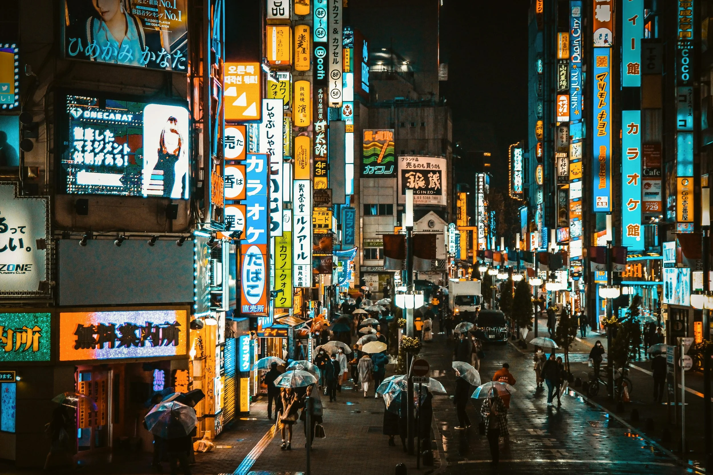 Narrow street at night lined with colorful neon signs and karaoke billboards, with people walking under umbrellas, Tokyo