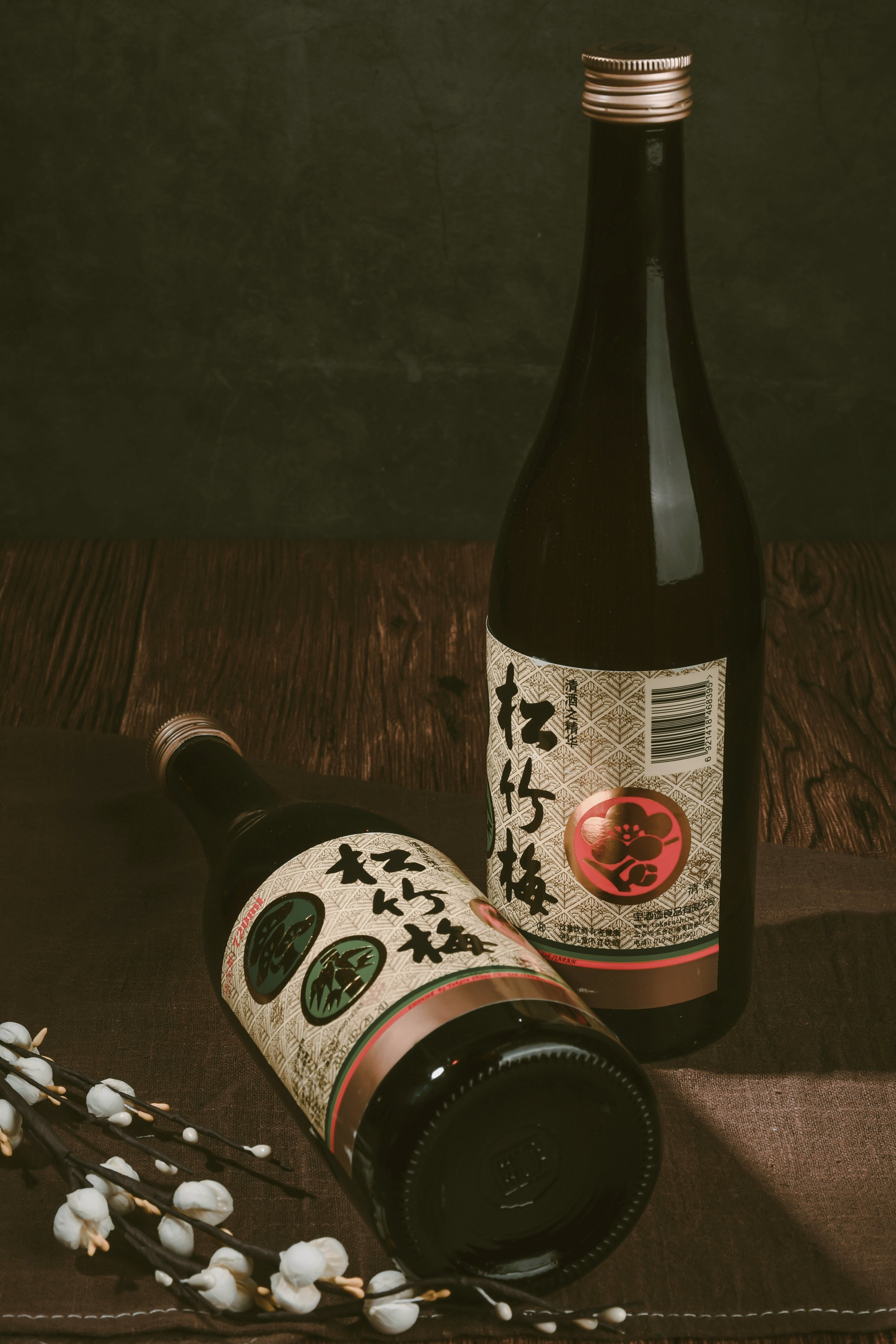 Two sake bottles against a dark background, one lying on its side beside small white flowers, Tokyo.