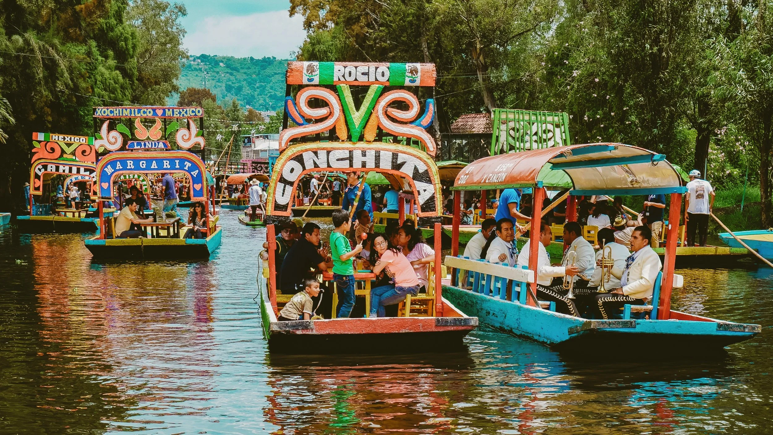 Colorful trajineras carrying groups of people along a canal lined with trees, Xochimilco, Mexico City.