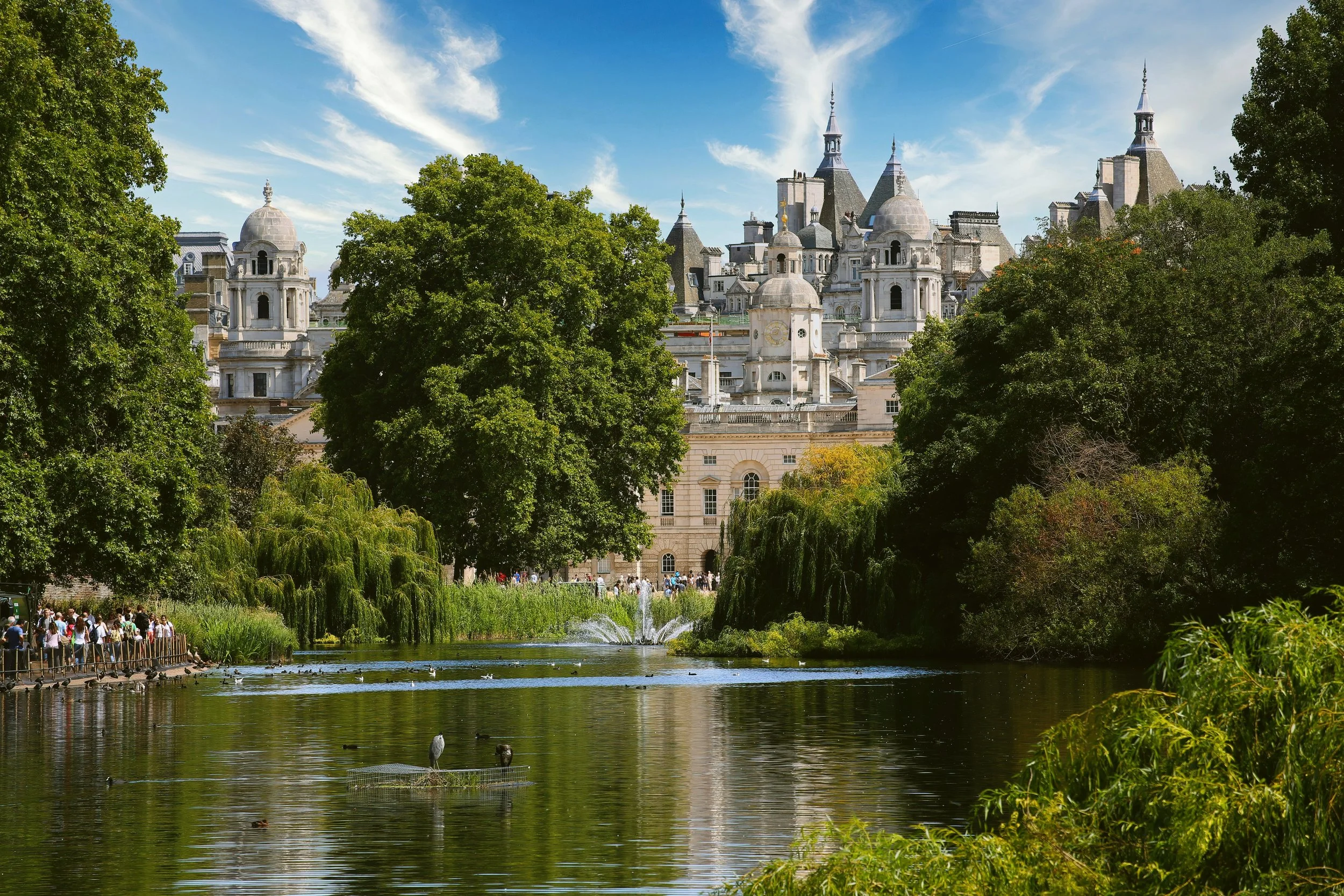 People rowing boats on the lake in St. James's Park, with Buckingham Palace visible among the trees