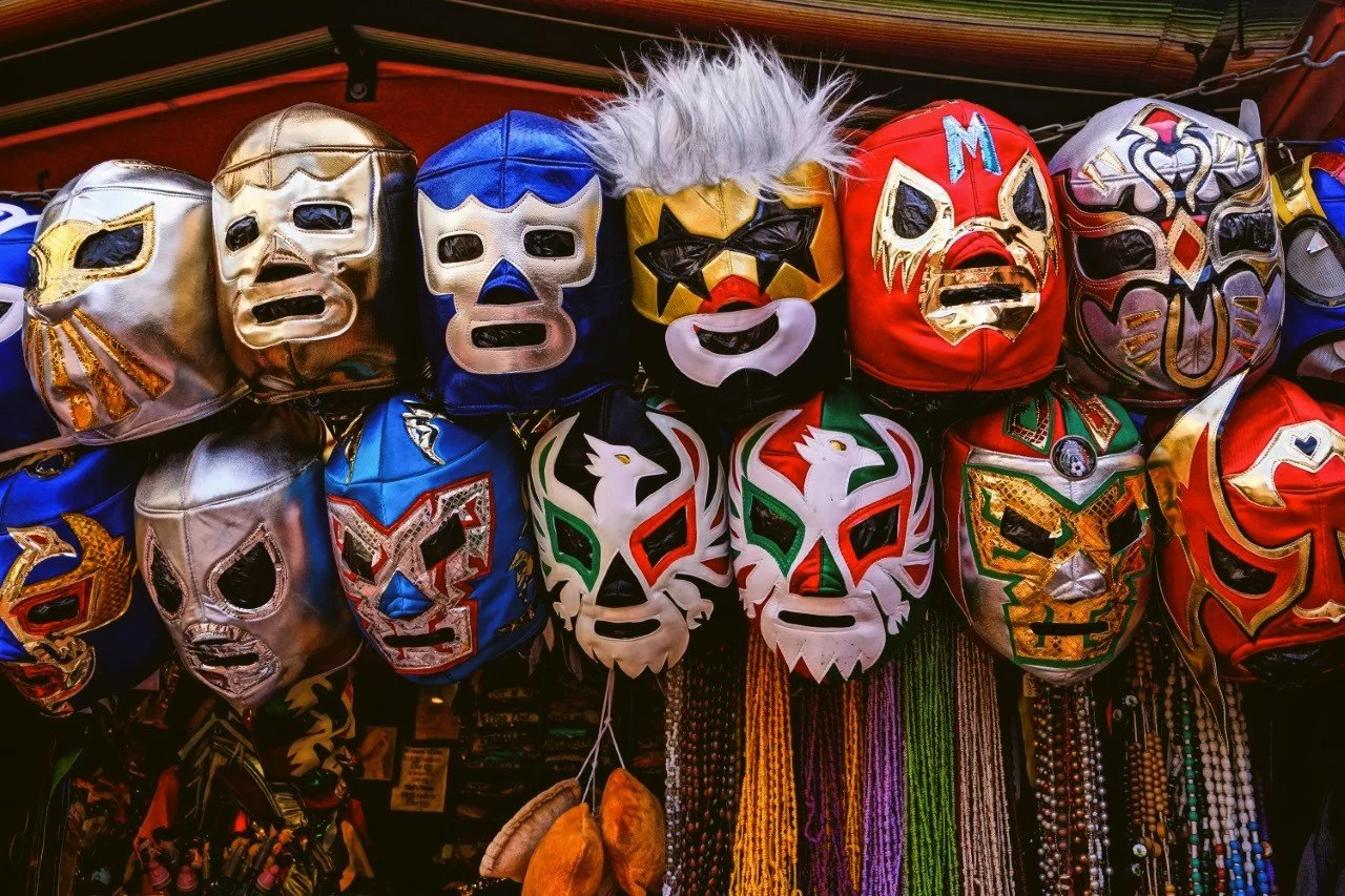 A display of colorful luchador masks hanging in a market stall, with beaded necklaces below, Mexico City.