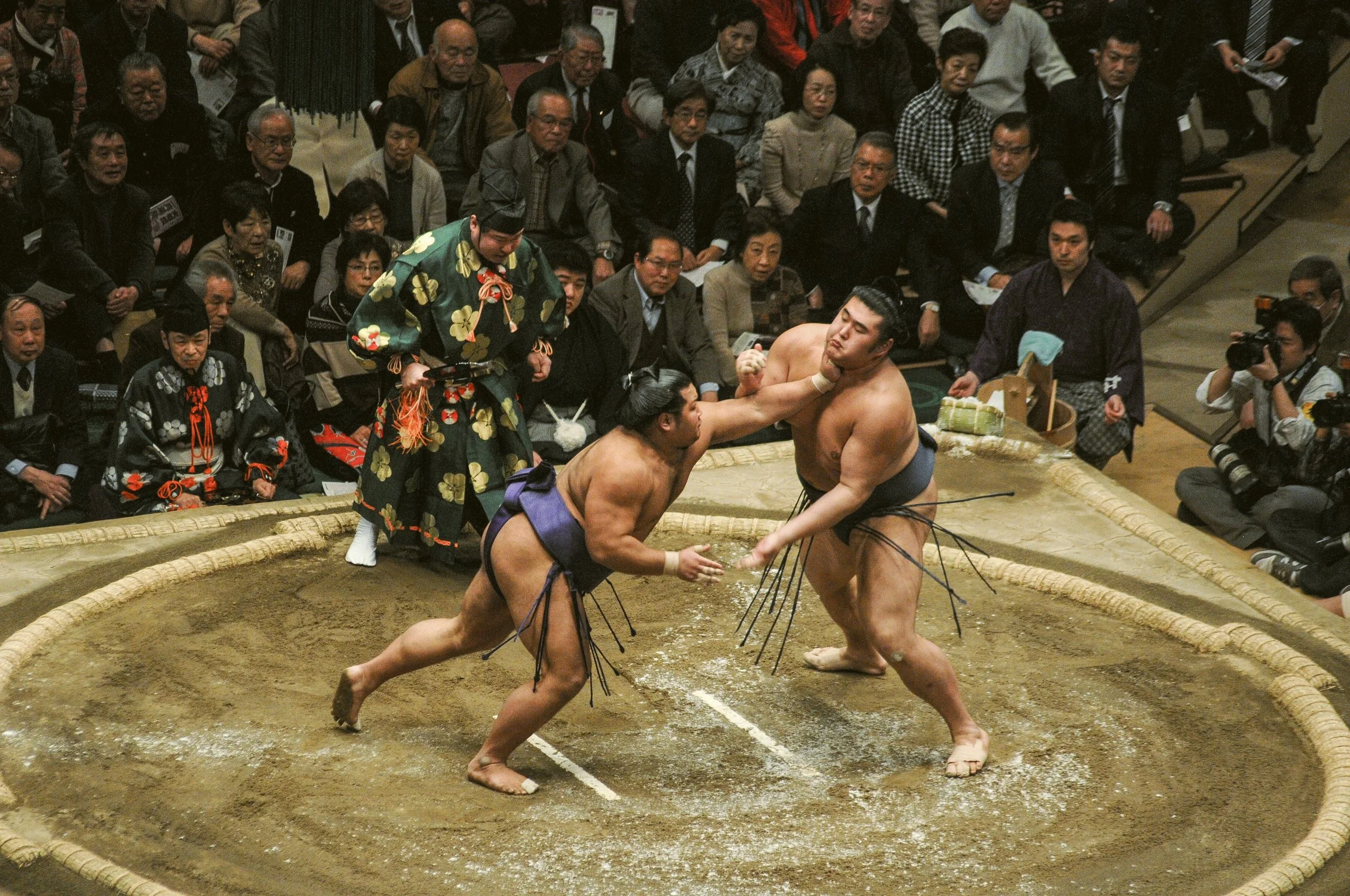 Two sumo wrestlers engaged in a match inside a sumo ring surrounded by a crowd of spectators. The wrestlers are in a stance, grappling with each other, while a referee in traditional attire observes closely.