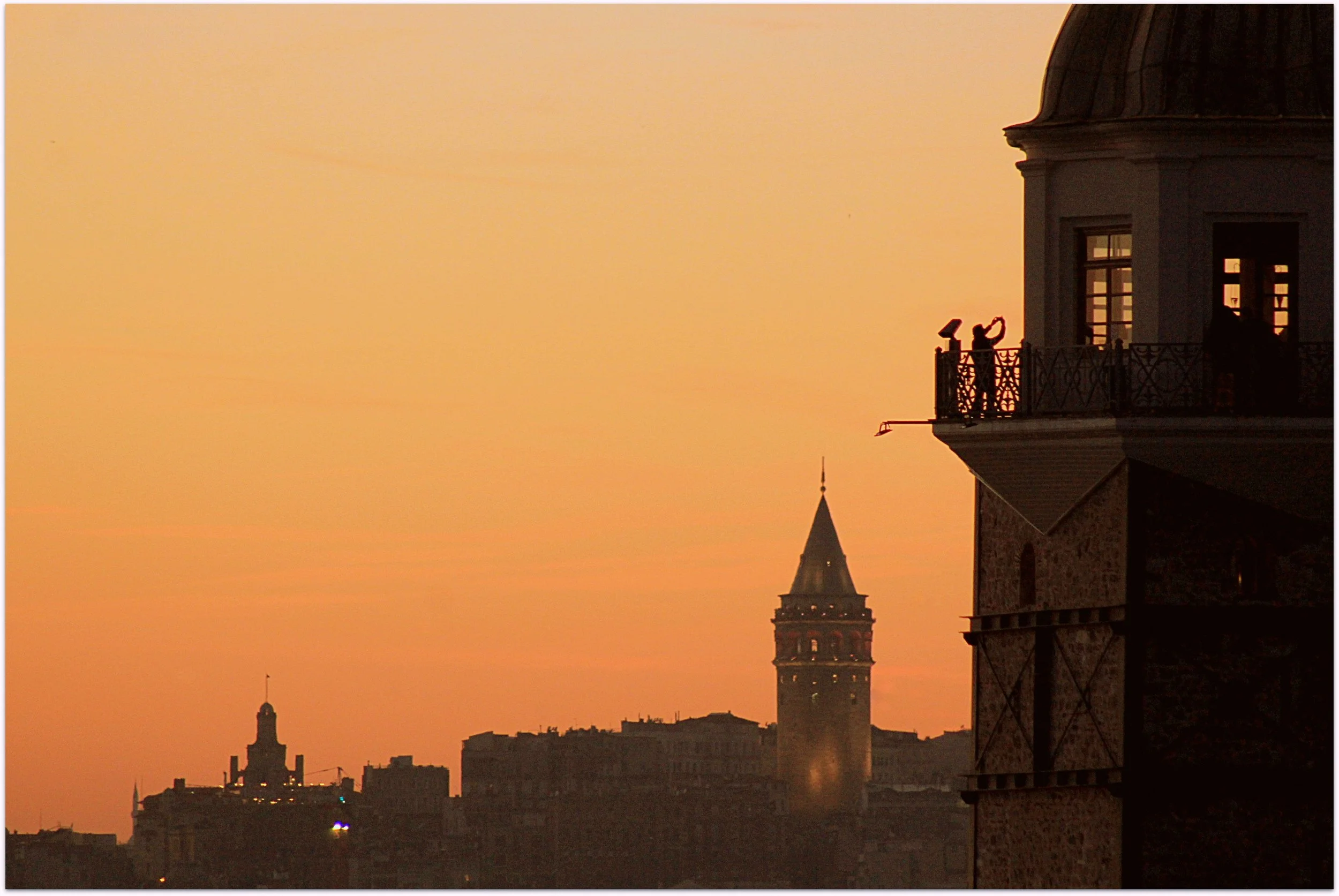 Galata Tower silhouetted against an orange sunset sky, with people gathered on a tower balcony in the foreground, Istanbul.