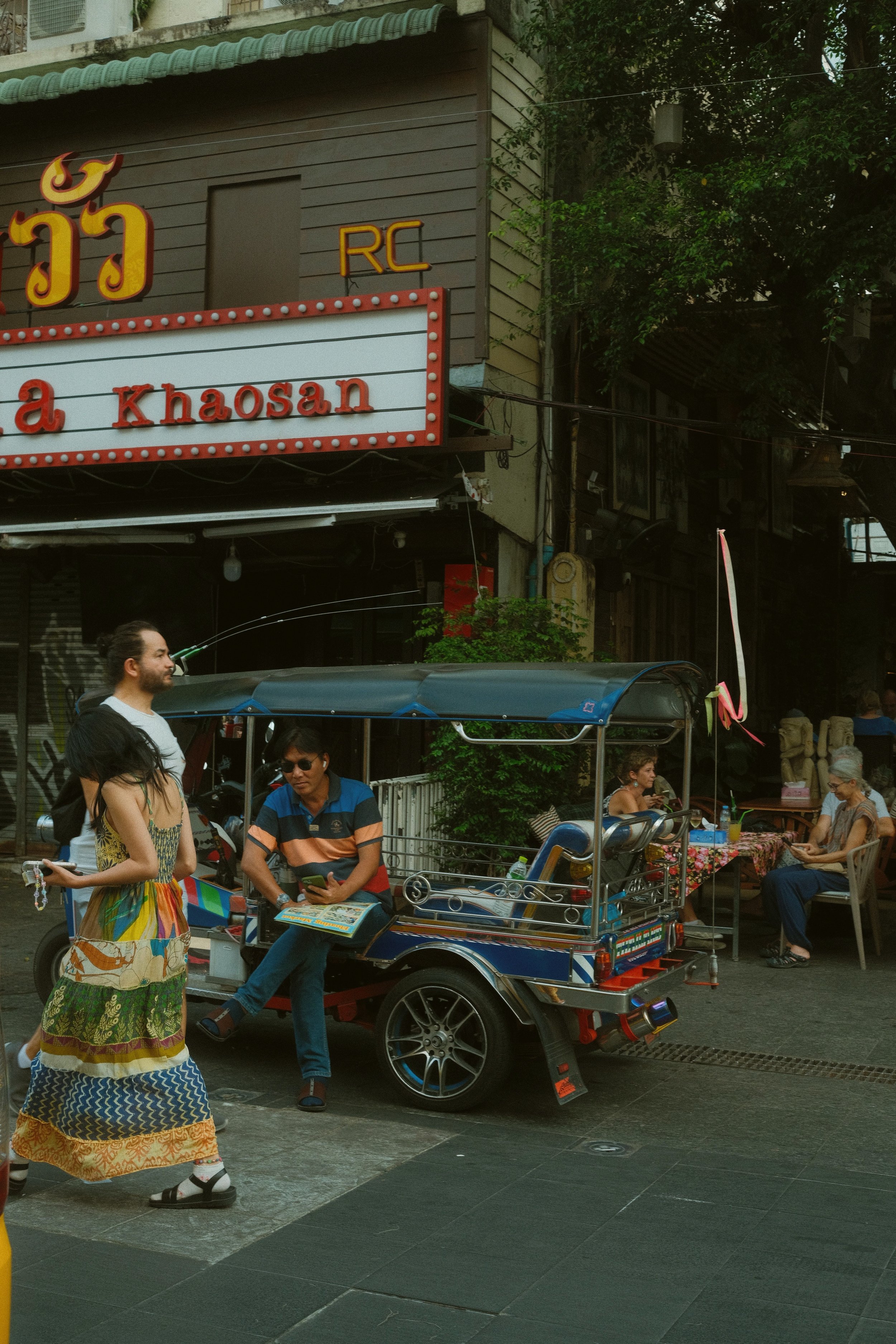 Street scene with a tuk-tuk and a golden statue in front of shops with colorful signs, Khao San Road, Bangkok.