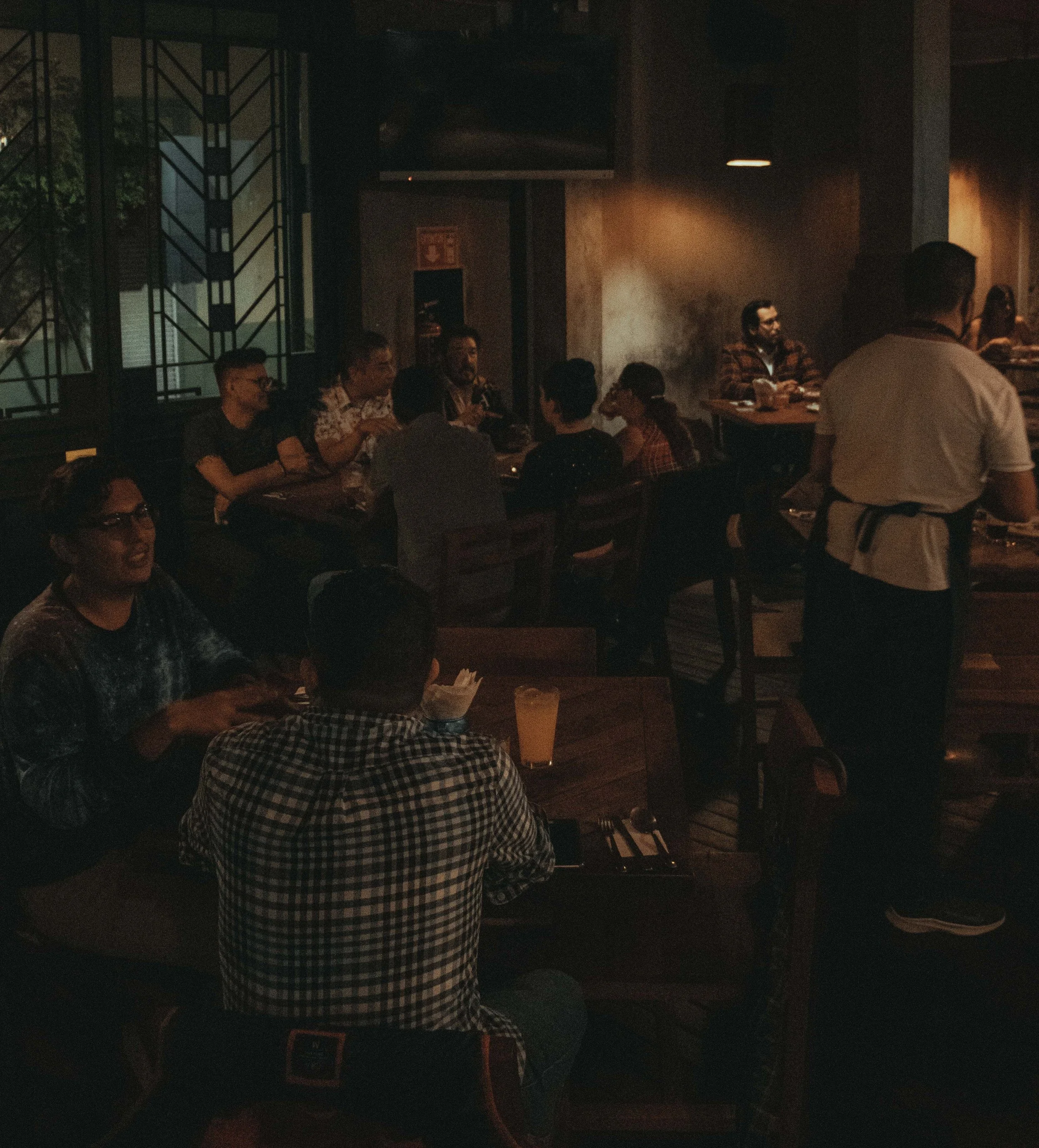 People sitting at tables in a dimly lit cantina, with some standing and engaging in conversation, Mexico City.
