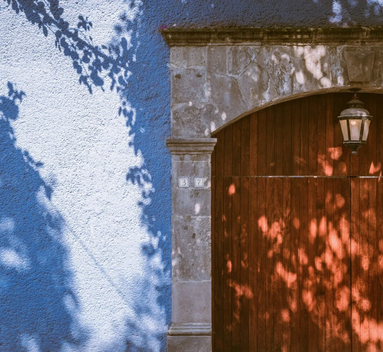 Reddish wooden door with a hanging lantern set in a stone doorway beside a blue wall with dappled shadows, Mexico City.