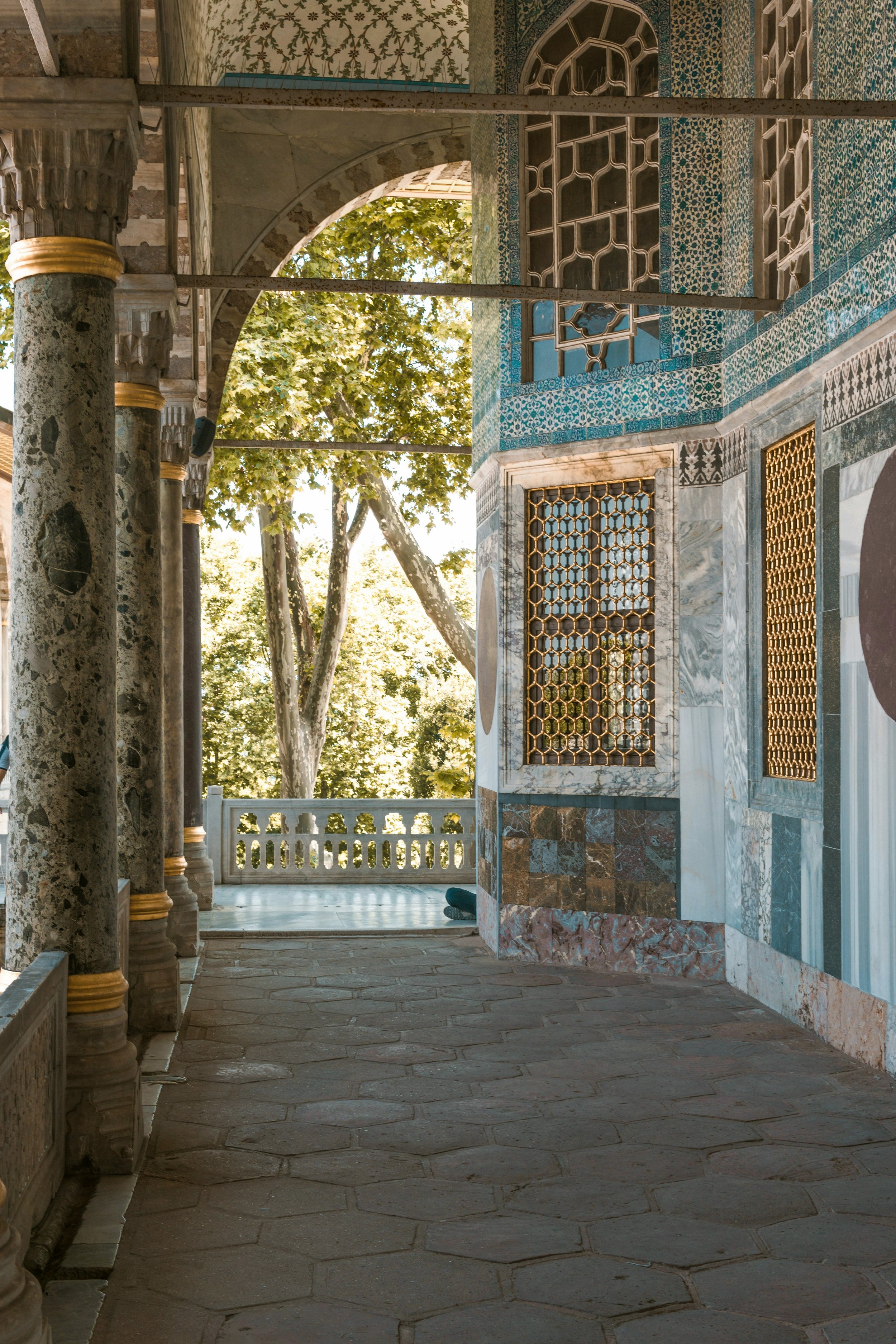 Stone gallery with arched openings looking onto a green courtyard and blue-tiled walls, Topkapı Palace, Istanbul.