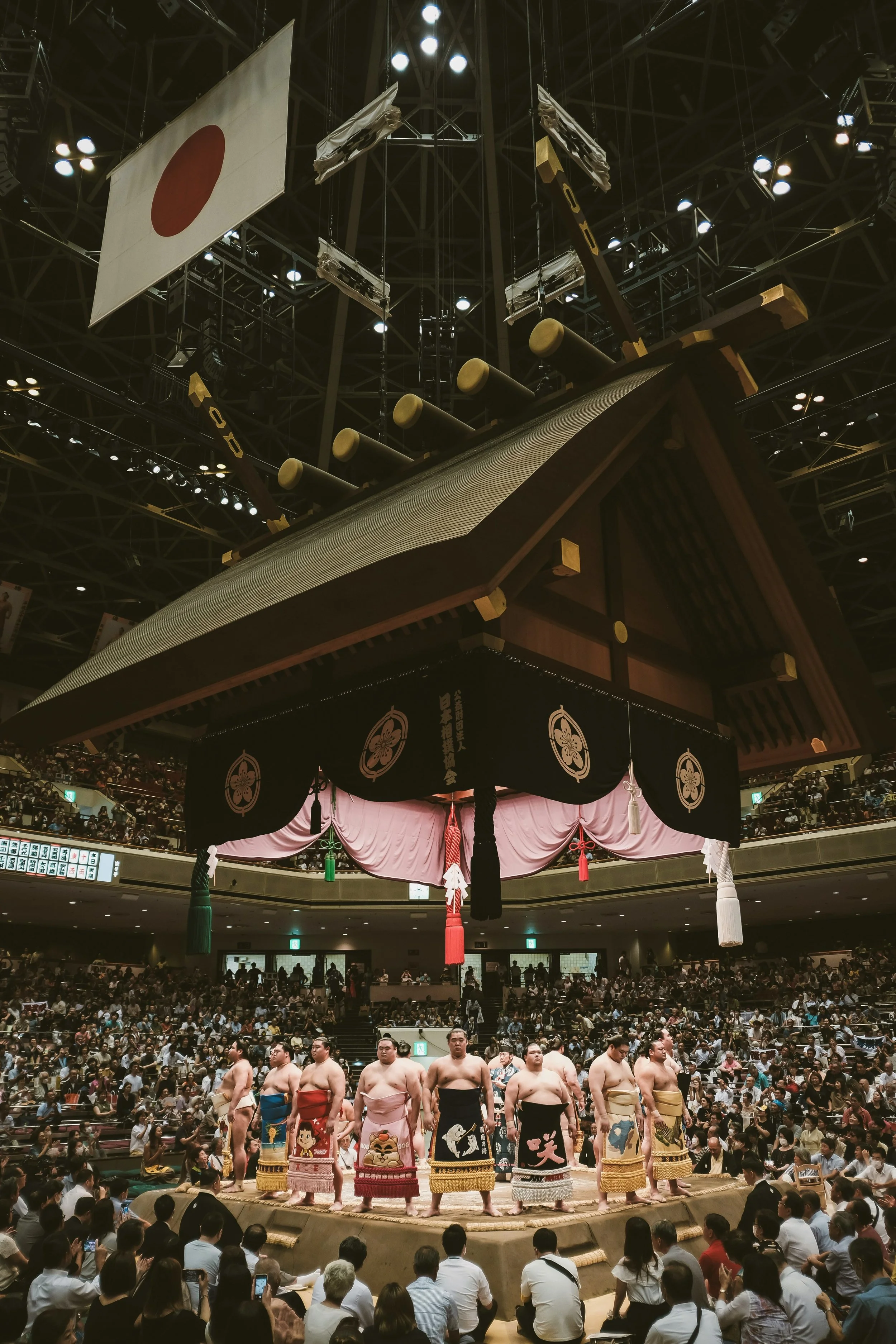 Sumo wrestlers standing in a row inside a sumo ring, with spectators watching from all sides of an indoor arena. A traditional large umbrella-like structure hangs above the ring, decorated with Japanese symbols, Tokyo.