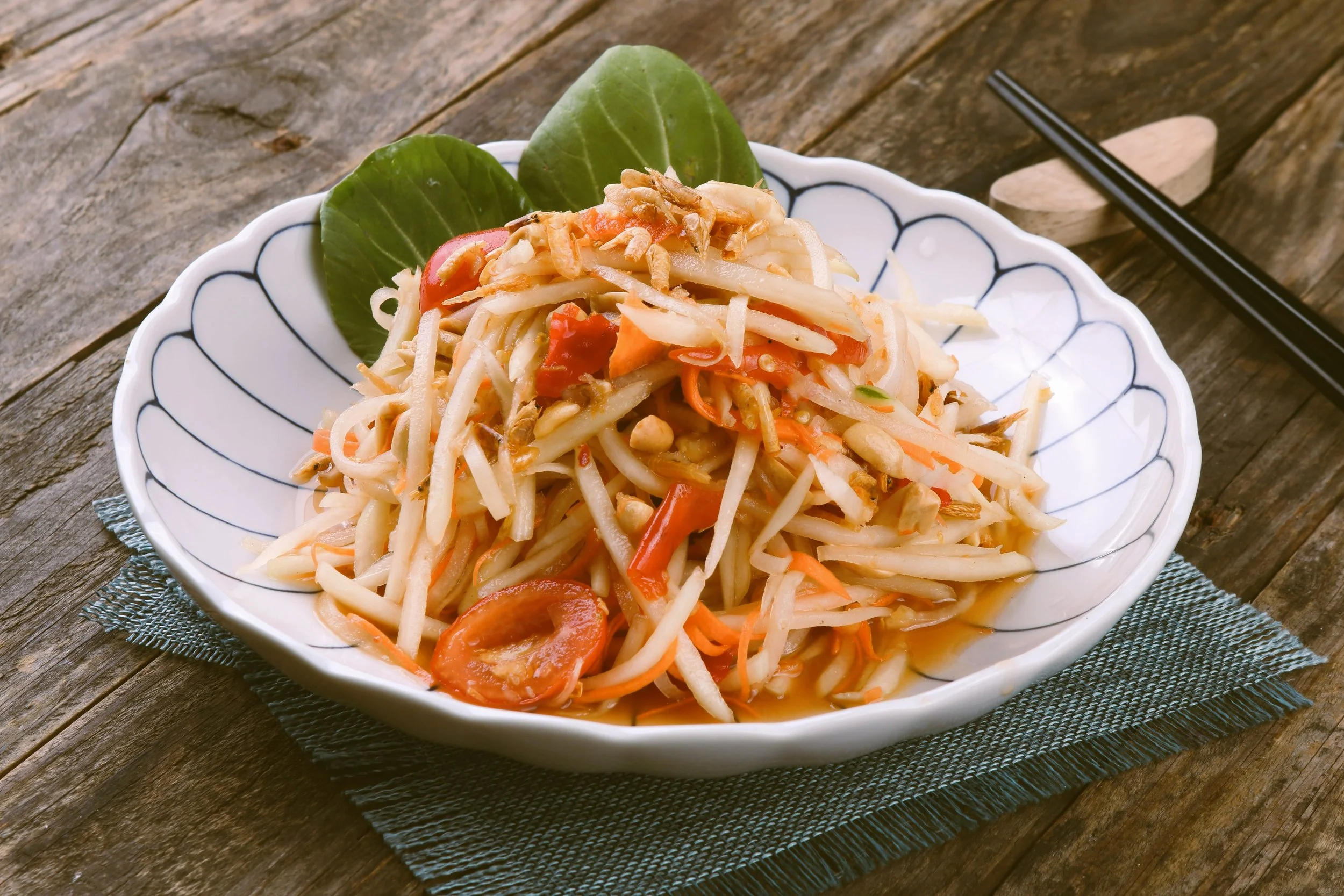 Bowl of som tam green papaya salad with shredded papaya and carrot on a rustic wooden table, Bangkok.