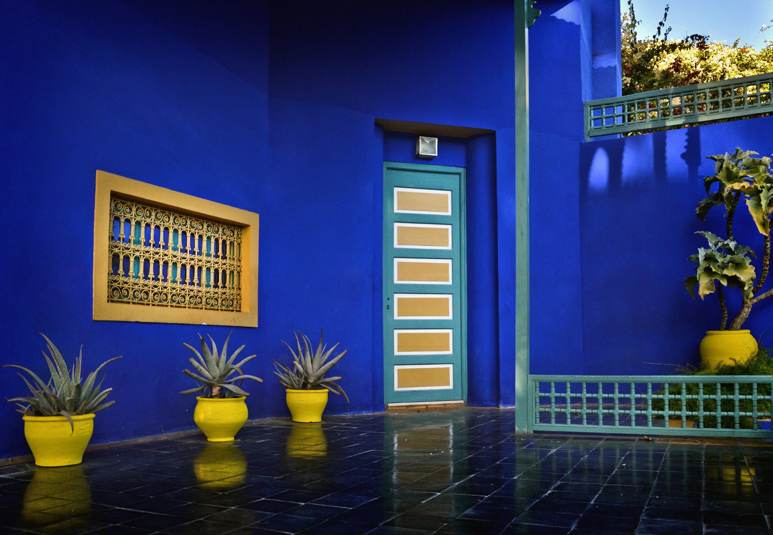 Cobalt-blue wall with a wooden window, a turquoise door and yellow pots reflected on the floor, Jardin Majorelle, Marrakech.