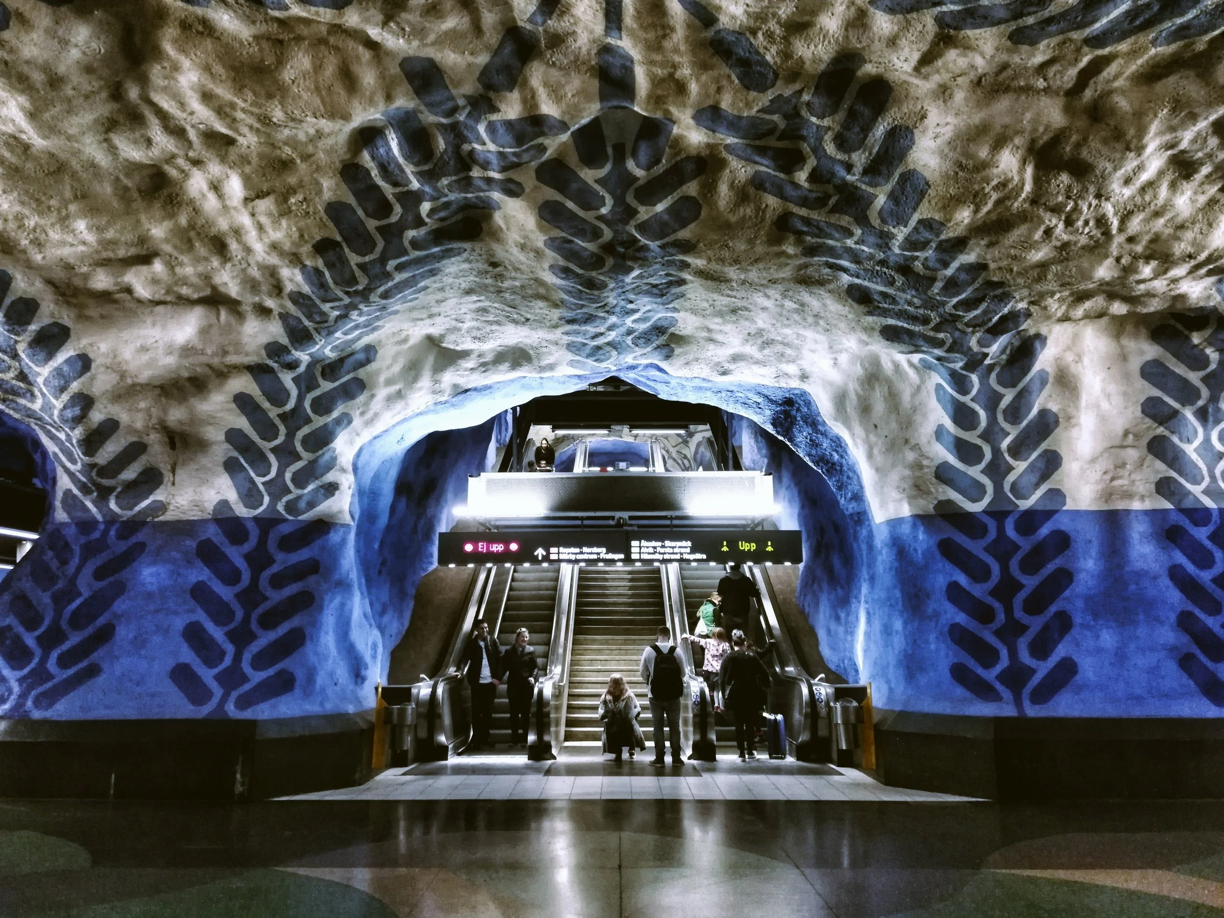 People riding escalators in a Stockholm metro station with decorative cave-like walls painted with blue and black patterns