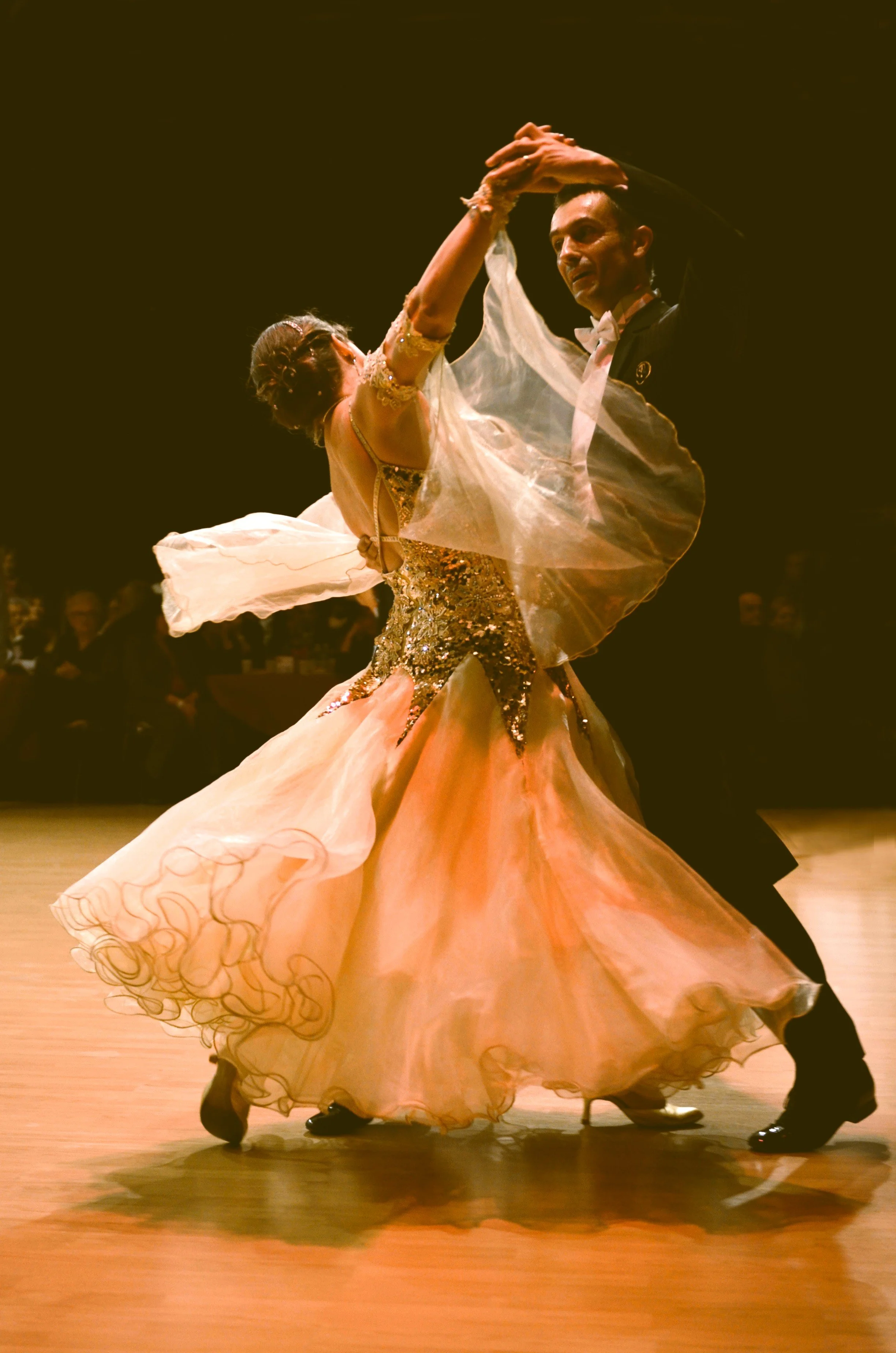 A couple dancing ballroom in formal attire, the woman in a flowing peach-colored gown with intricate gold details and the man in a black tuxedo, on a wooden dance floor with a dark background and an audience in the distance.