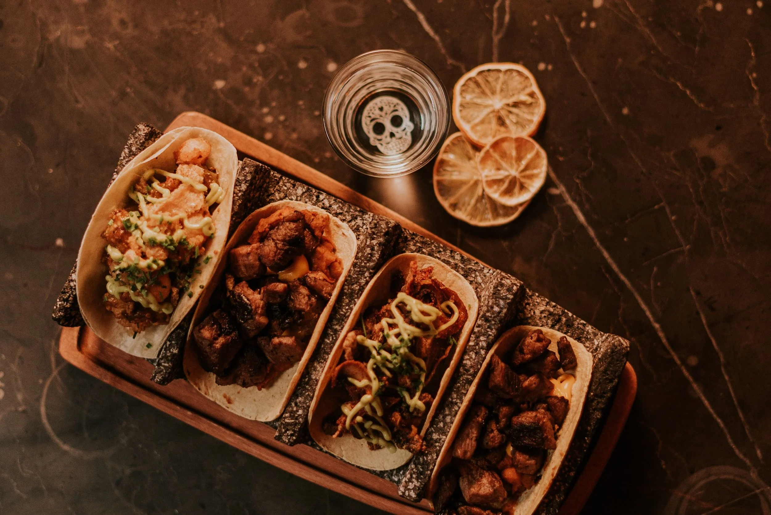 A wooden tray holding several tacos with various meats and toppings, in front of a glass of water with a skull design, and dried lemon slices on a dark marble surface.