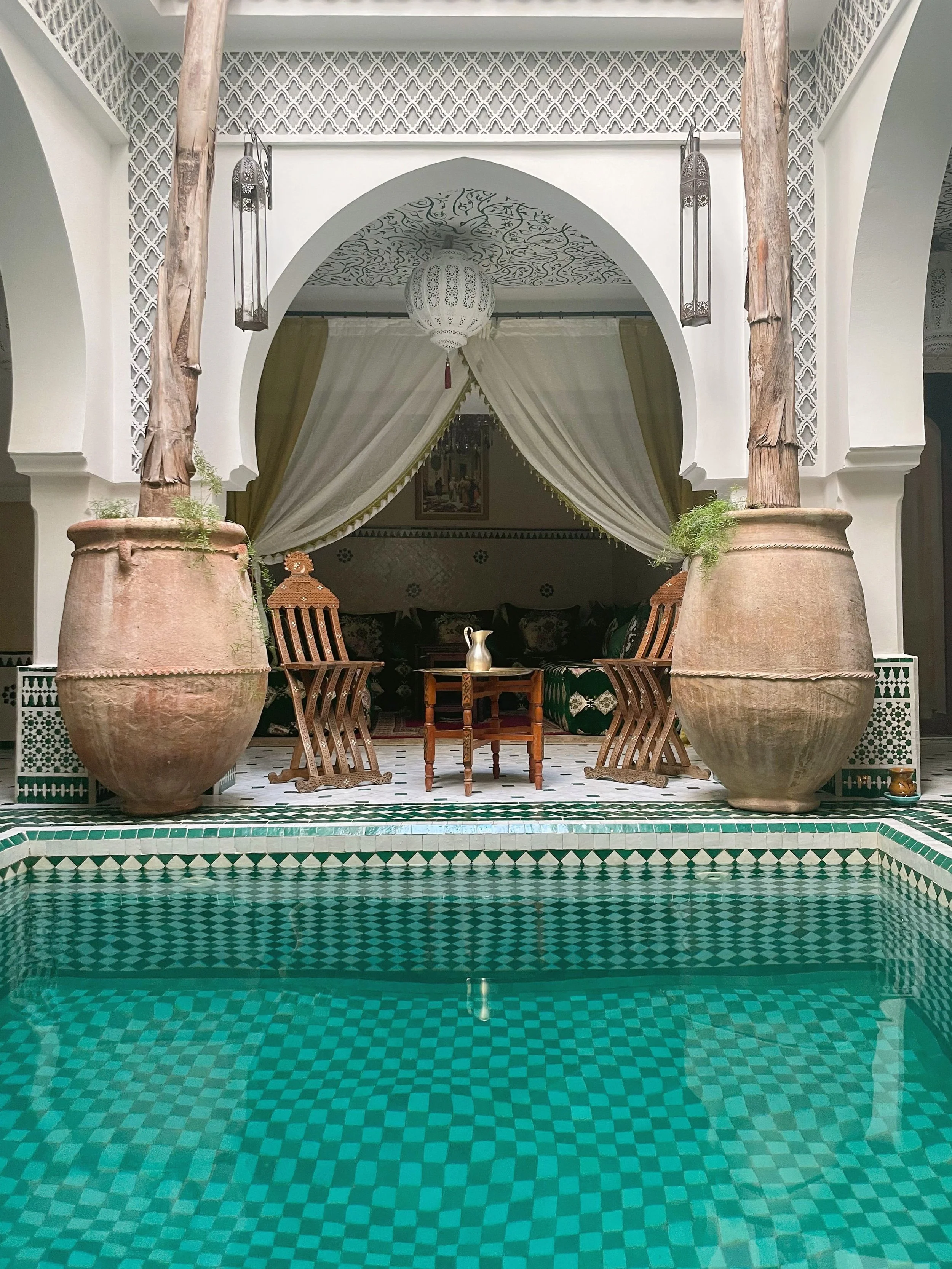 Riad courtyard with a turquoise pool, large clay urns and a lounge area under a draped canopy, Marrakech.
