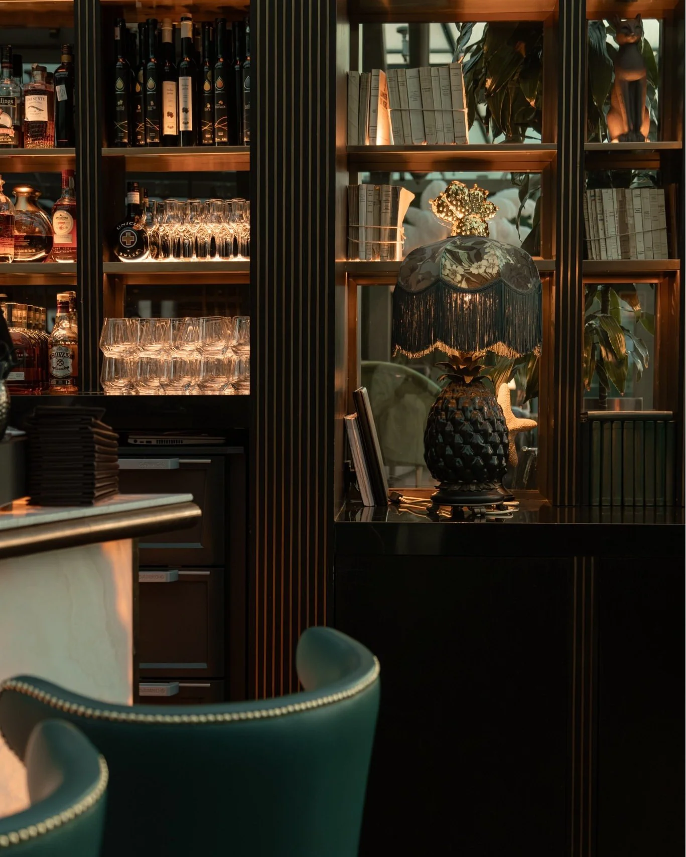 A shelf room with books, glasses, and liquor bottles behind a well-lit area. There's a vintage lamp with a floral lampshade and a pineapple-shaped base on a black counter in front of the shelf, along with a green chair in the foreground.