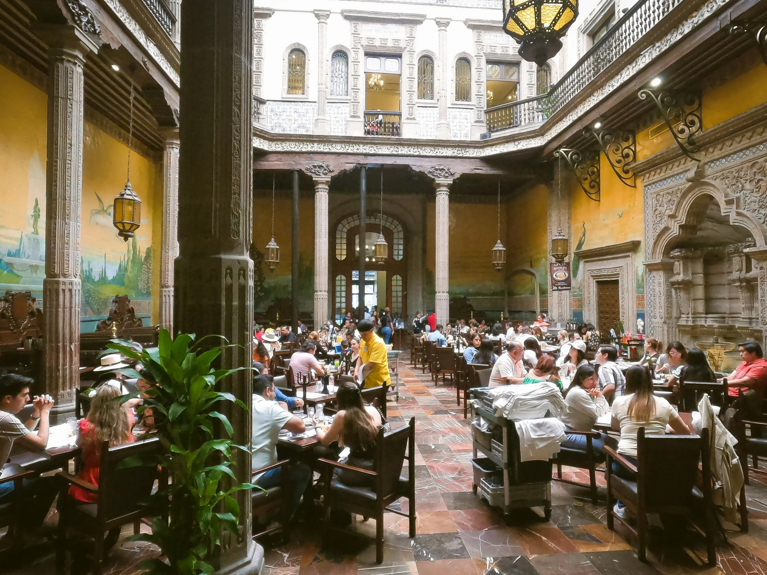 Diners seated at tables in the colonial courtyard of Casa de los Azulejos, with columns, ochre walls and plants, Mexico City.