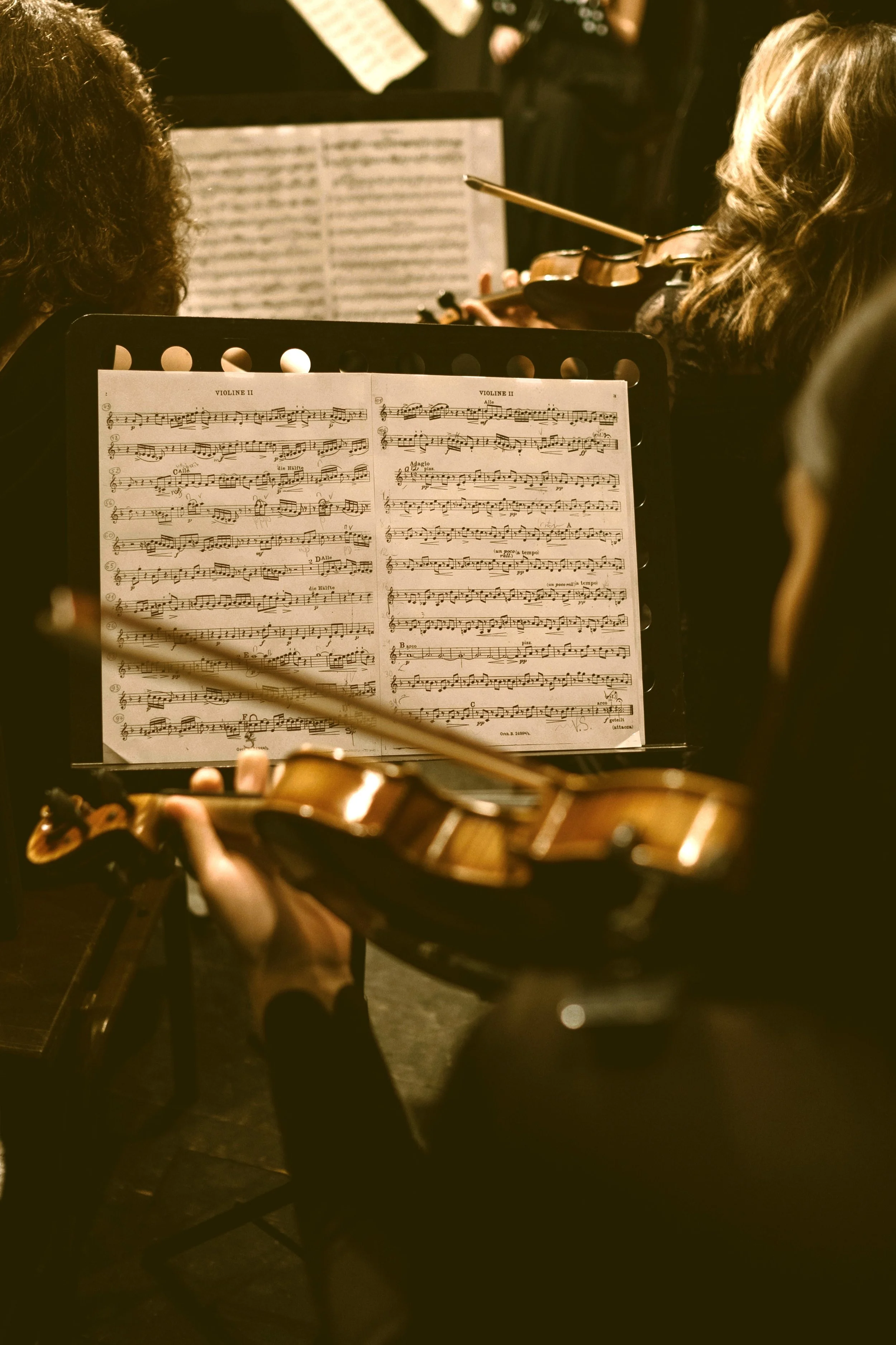 Close-up of music sheet on a stand, with a violin in the foreground and a person playing the violin in the background in a dimly lit room.
