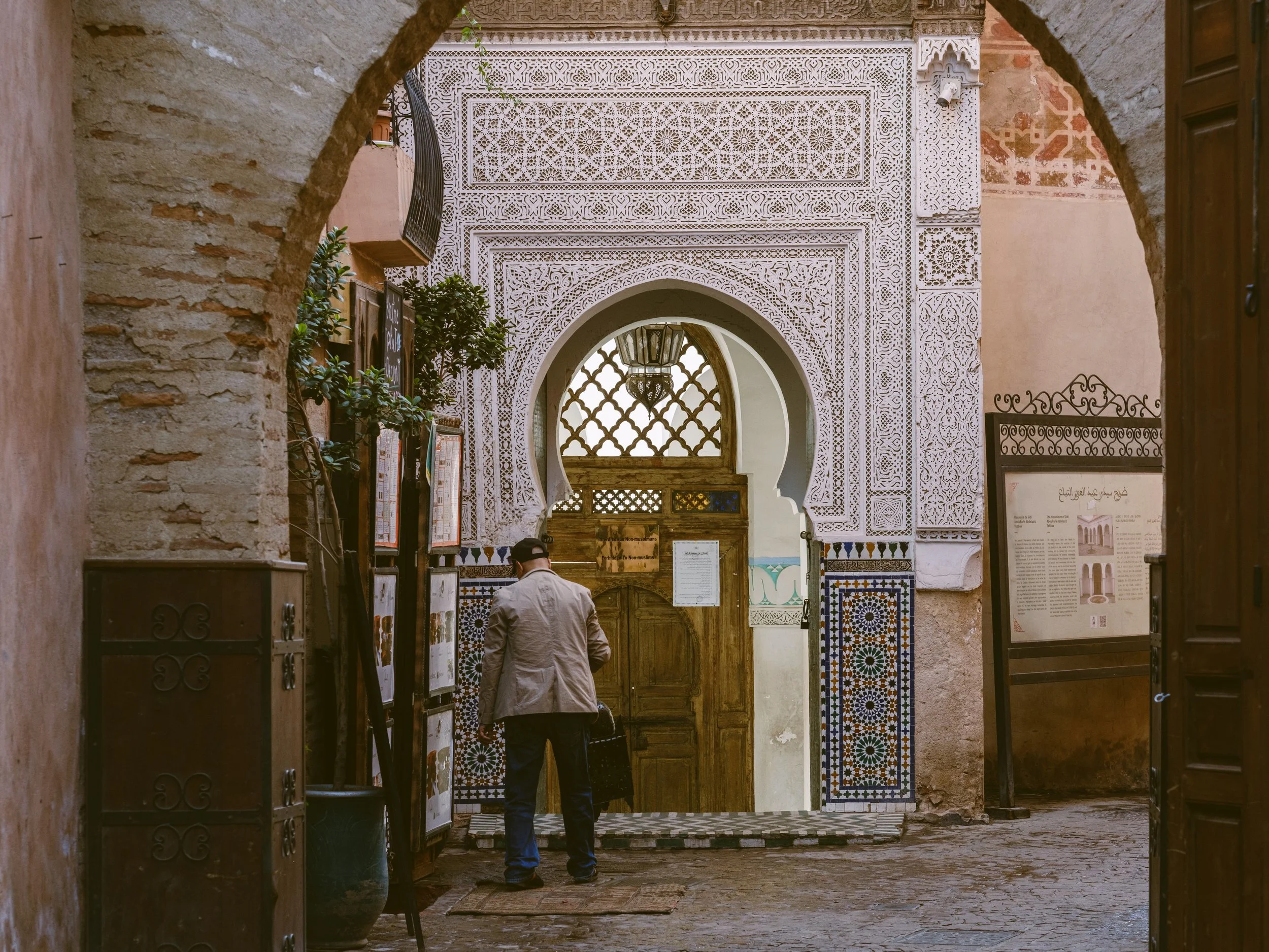 Person standing in a doorway framed by an arch, with an ornate carved stucco facade behind, Marrakech.