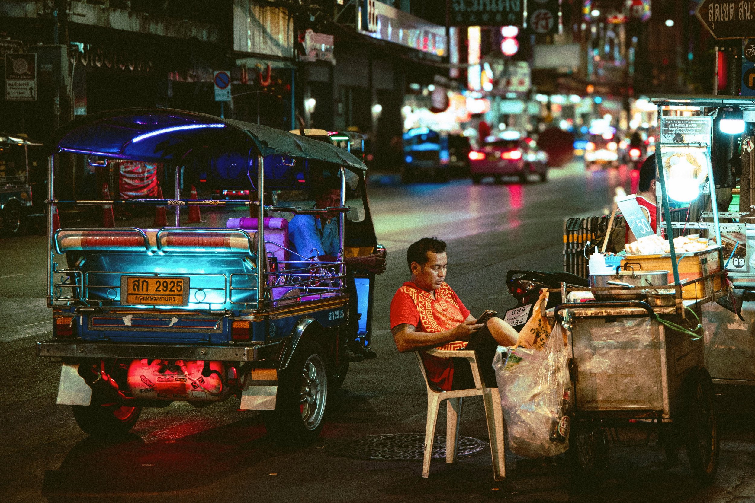 Neon-lit tuk-tuk parked on a dark street at night beside a street vendor seated on a plastic stool, Bangkok.