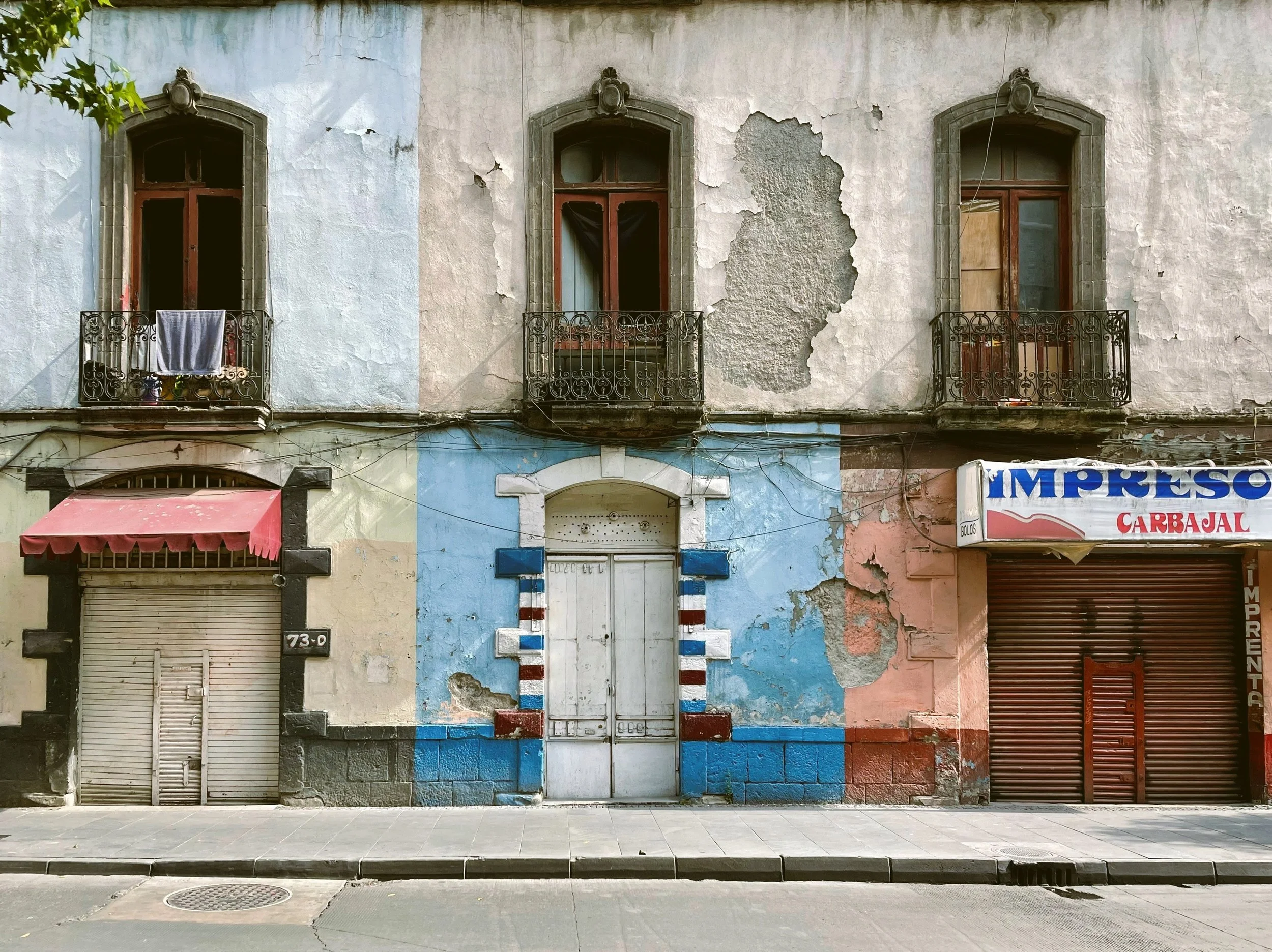 Weathered colonial facade with peeling blue and pink paint, arched windows and closed shop doors, Mexico City.