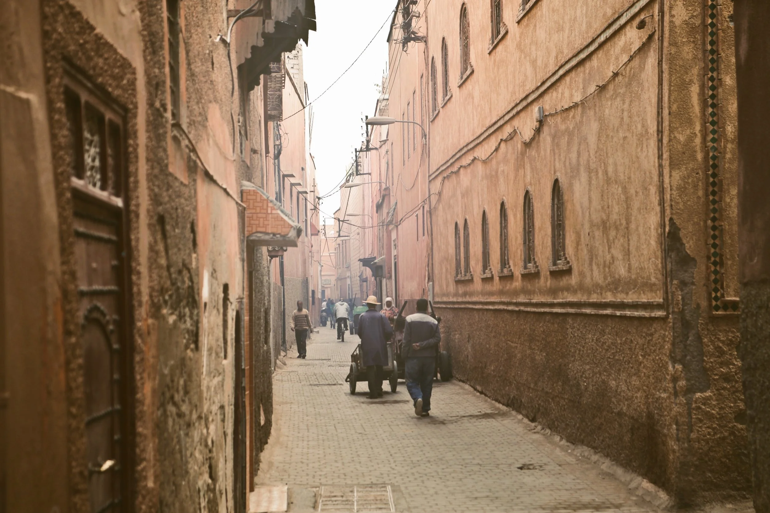 A narrow alleyway with old, textured buildings on either side. Several people are walking down the cobblestone street, and there are overhead wires, Marrakech.