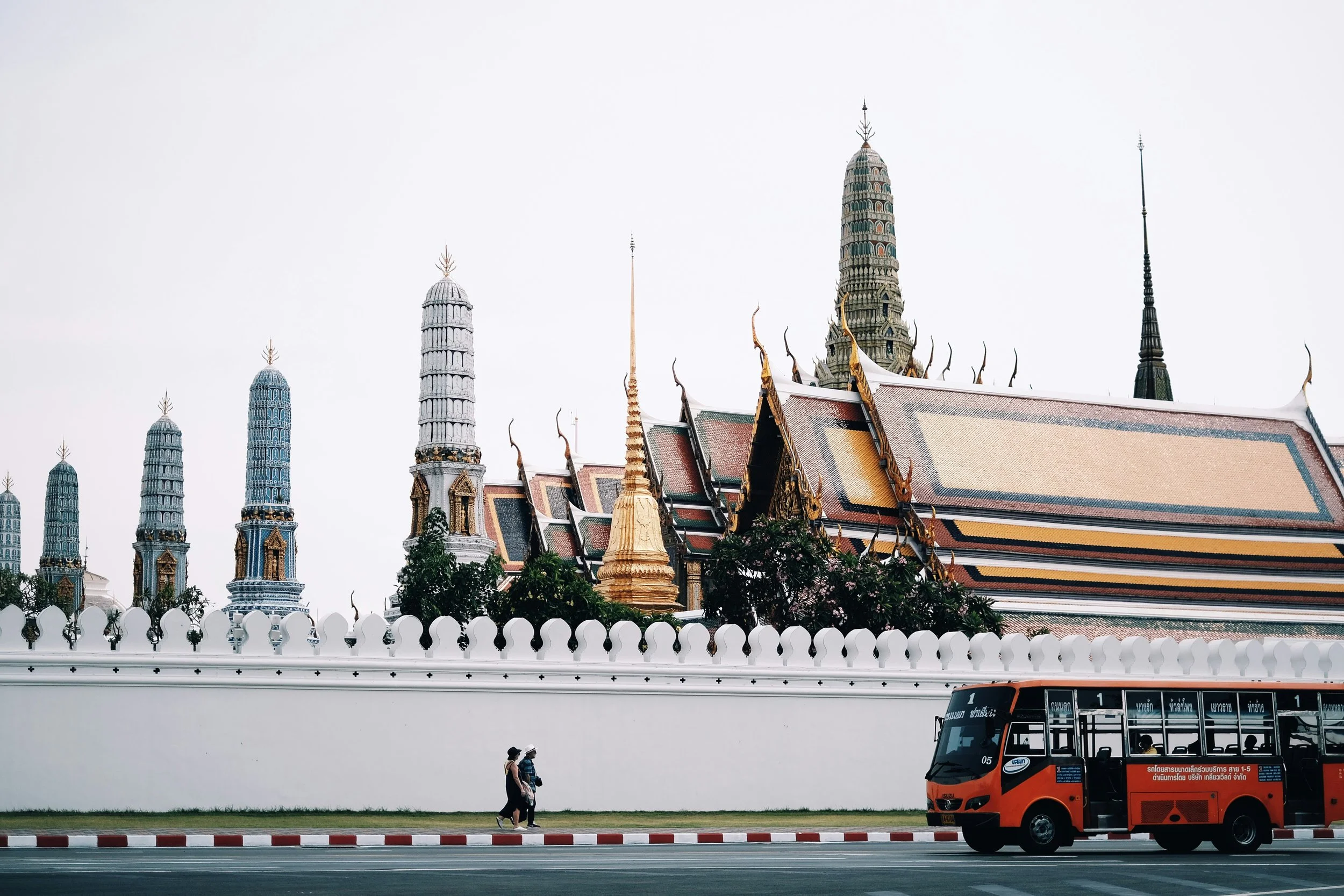 Grand Palace wall with golden spires and temple roofs behind it, with a red bus passing in front, Bangkok.