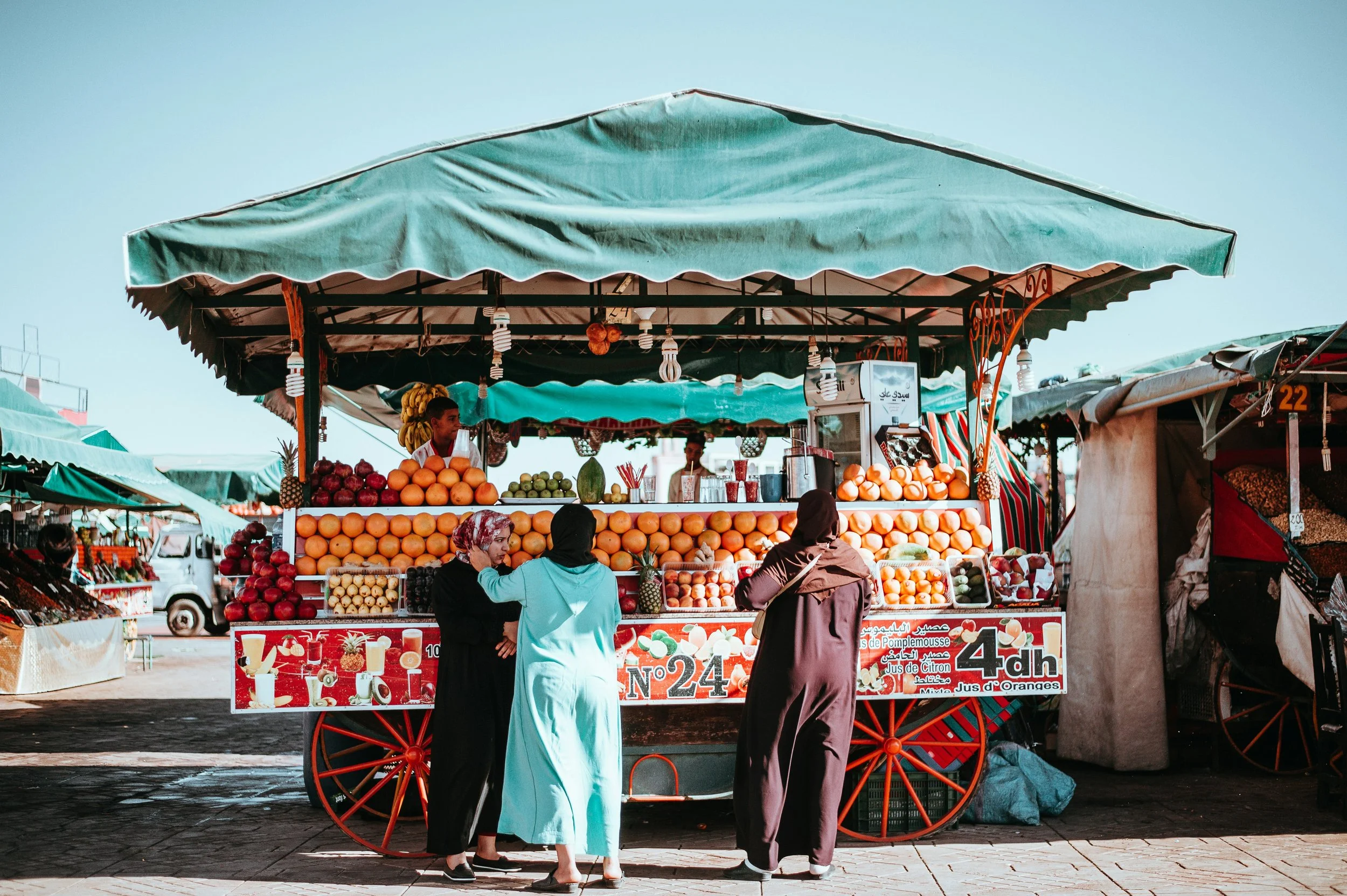 Orange juice stall with a green awning and customers standing at the counter, Marrakech.