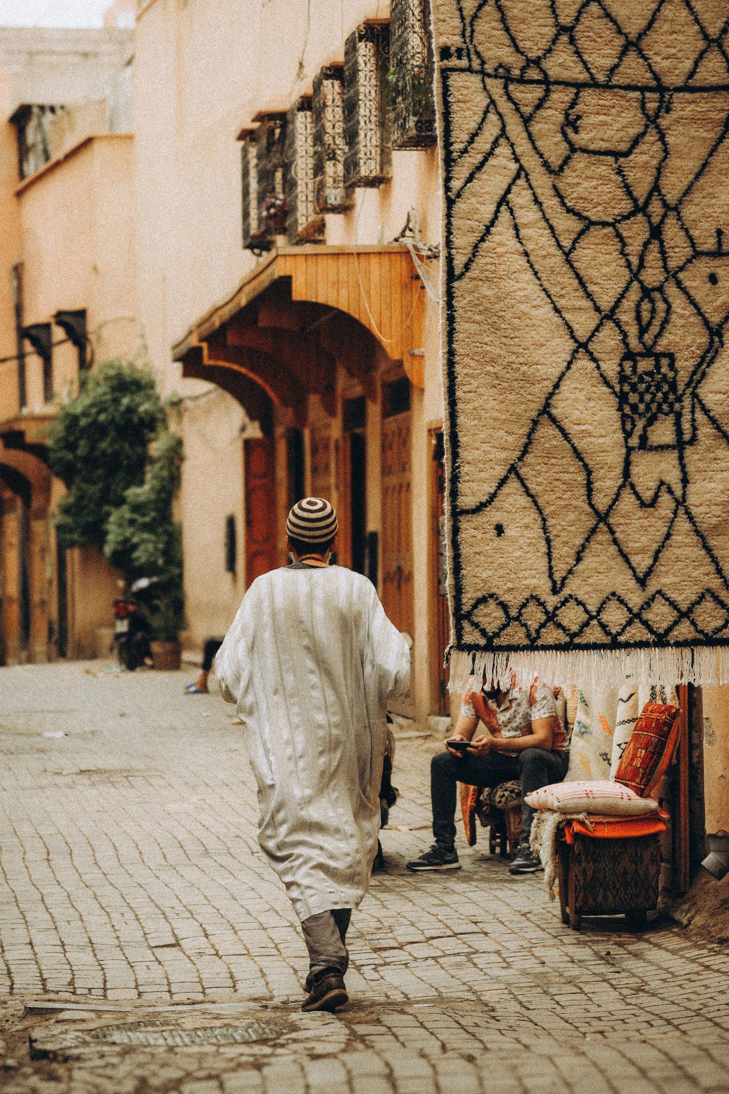 Man in a white djellaba walking along a sunlit medina alley with ochre walls, Marrakech.