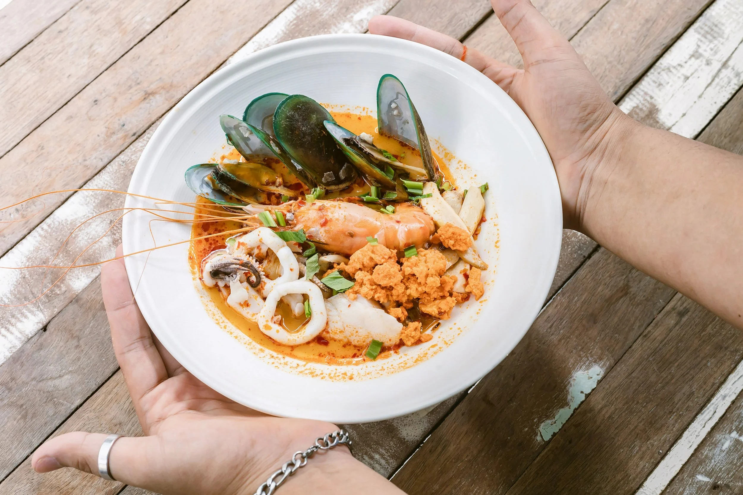 Hands holding a bowl of tom yum goong with shrimp, mussels and squid over a wooden table, Bangkok.