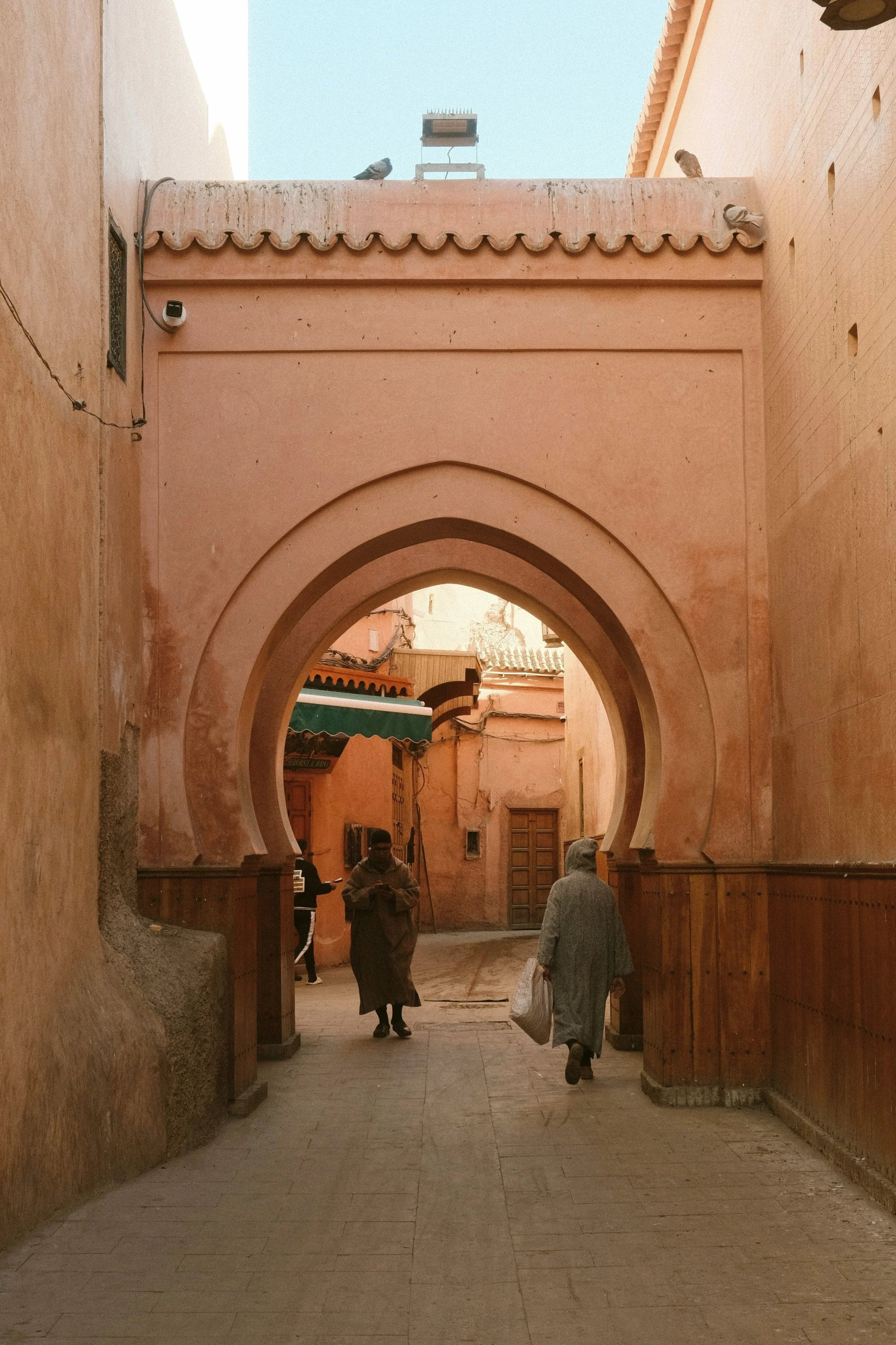 Narrow terracotta-colored alley with an arched passage and people walking through, Marrakech medina.