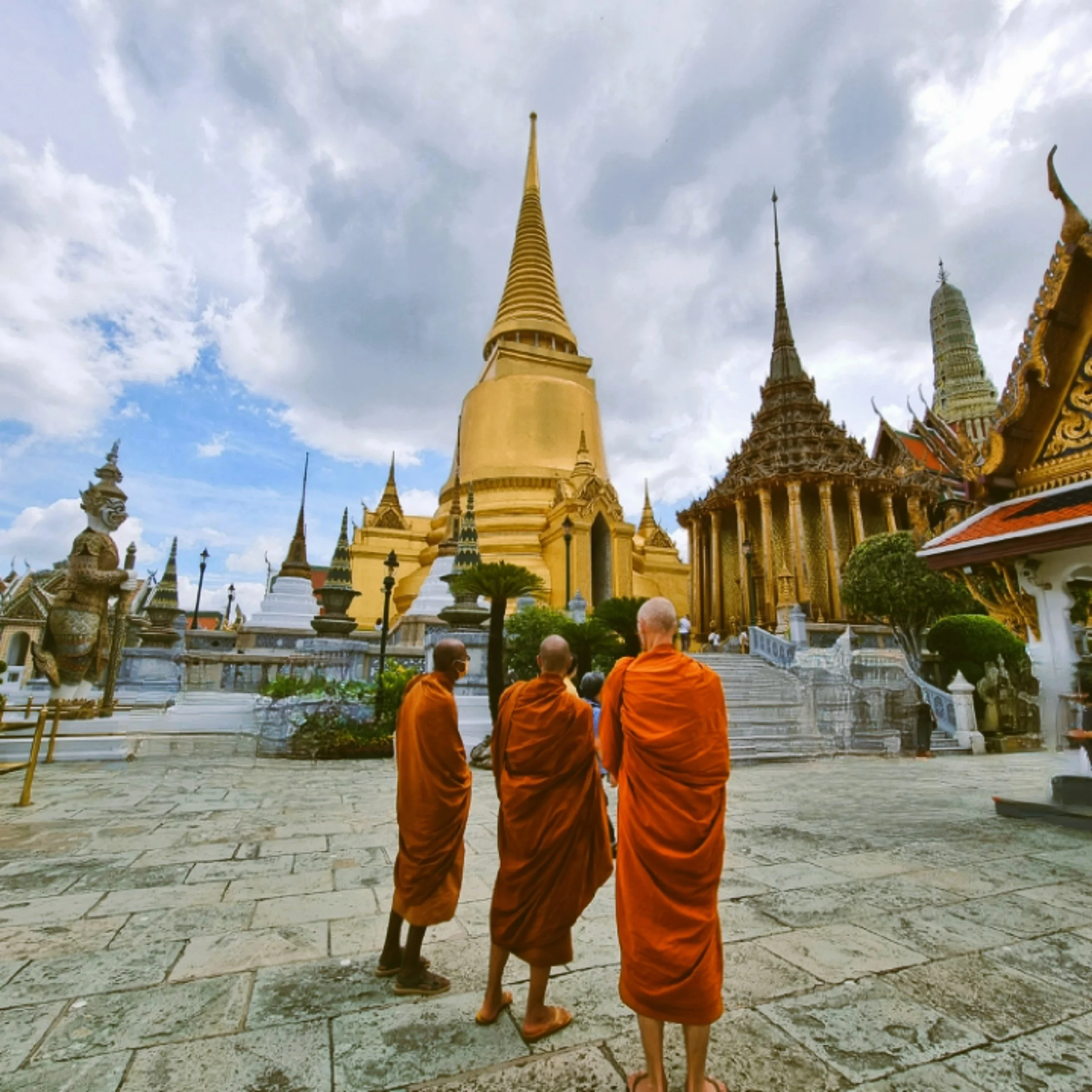 Three monks in orange robes walking across a stone courtyard toward a golden chedi, Grand Palace, Bangkok.