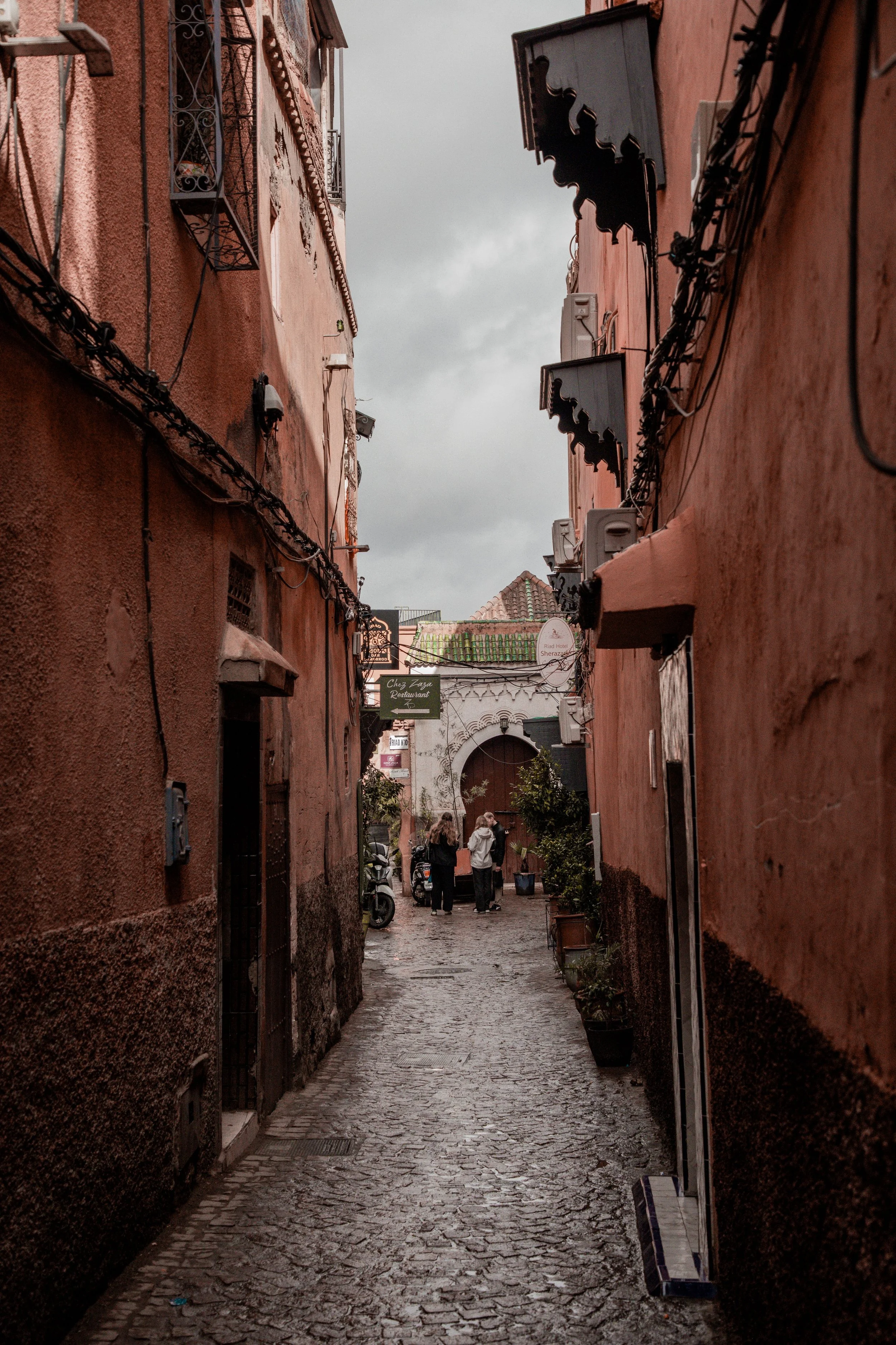Narrow medina alley with tall terracotta walls and a few people in the distance, Marrakech.