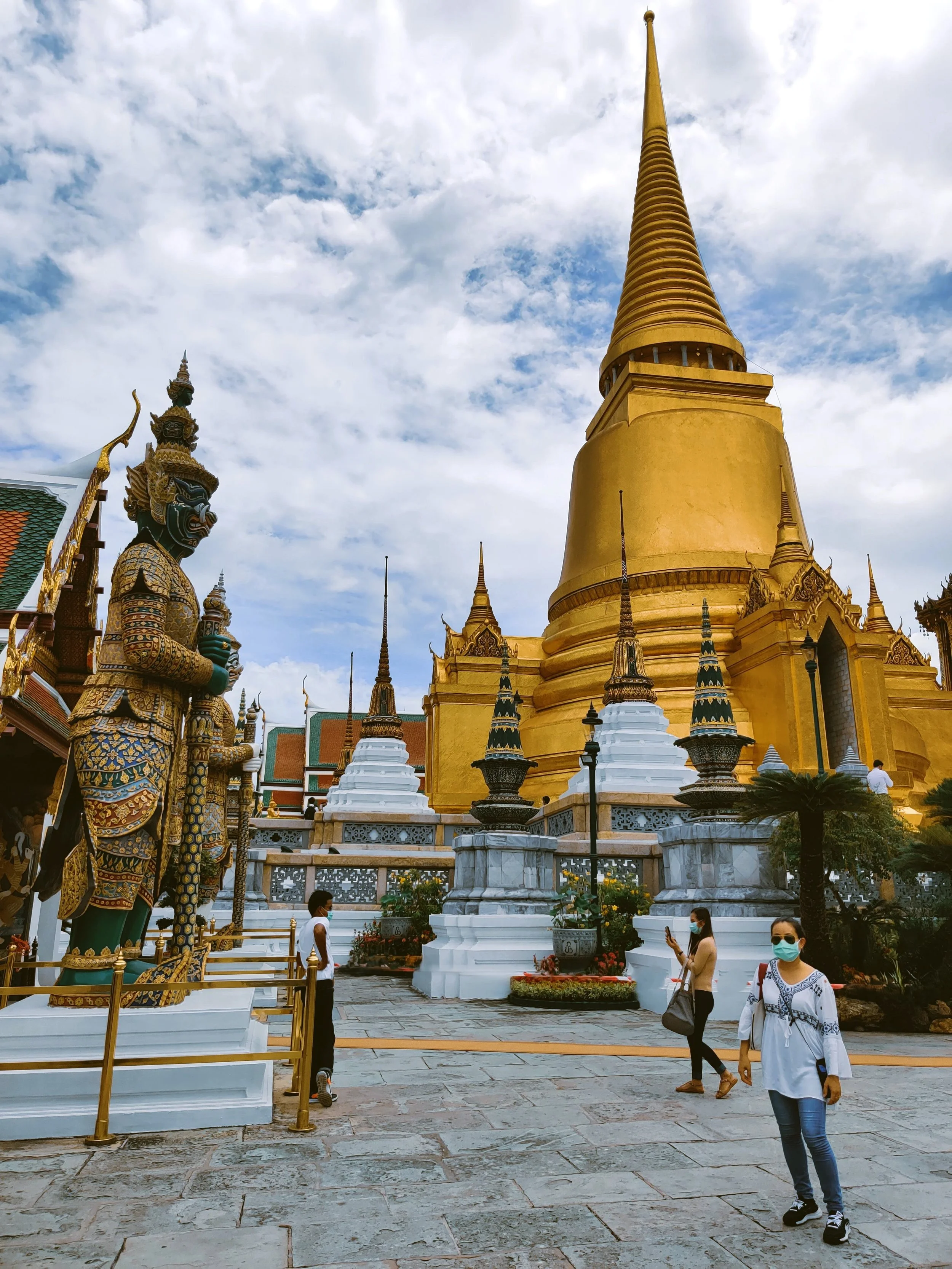 Golden chedi rising above a courtyard with a guardian statue on the left and visitors below, Grand Palace, Bangkok.