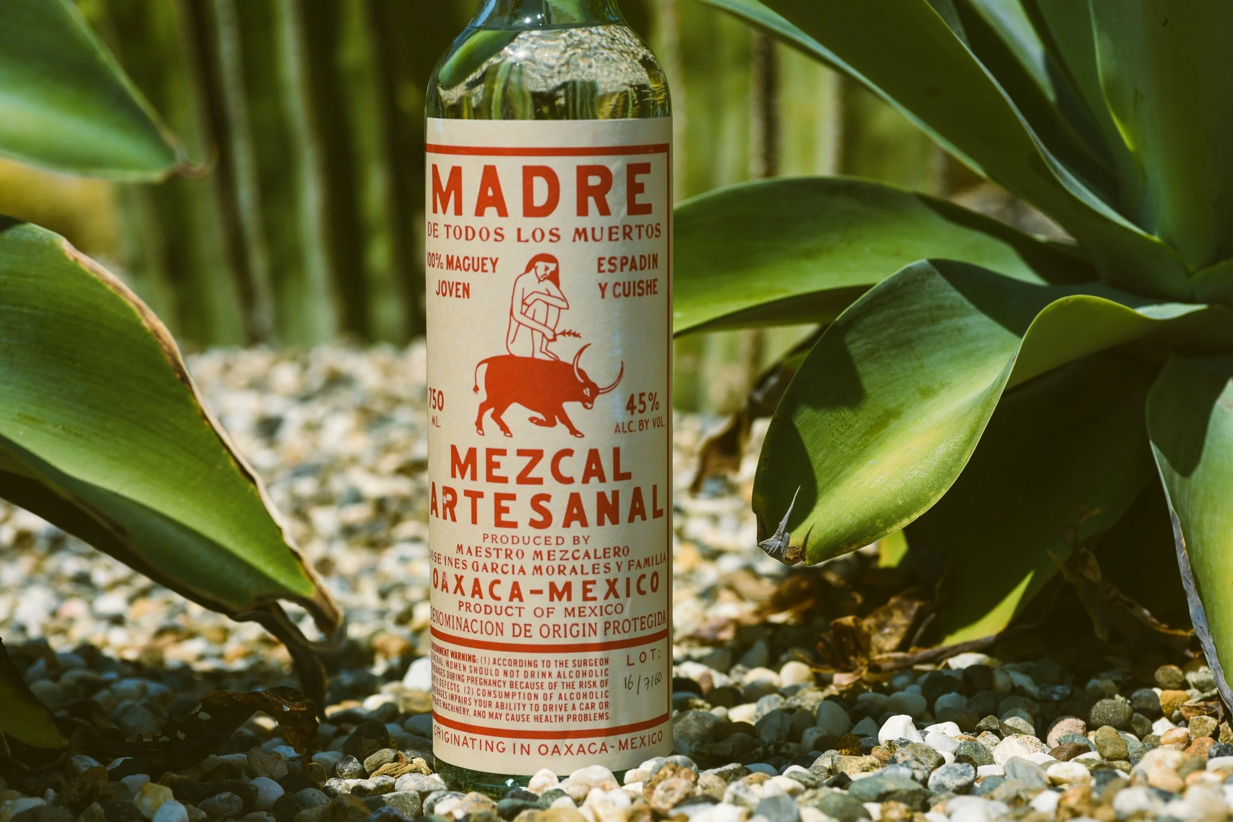 A bottle of Mexican mezcal with a red and white label that says 'Madre de todos los muertos' and features an illustration of a figure riding a goat, amidst green plants and small rocks.